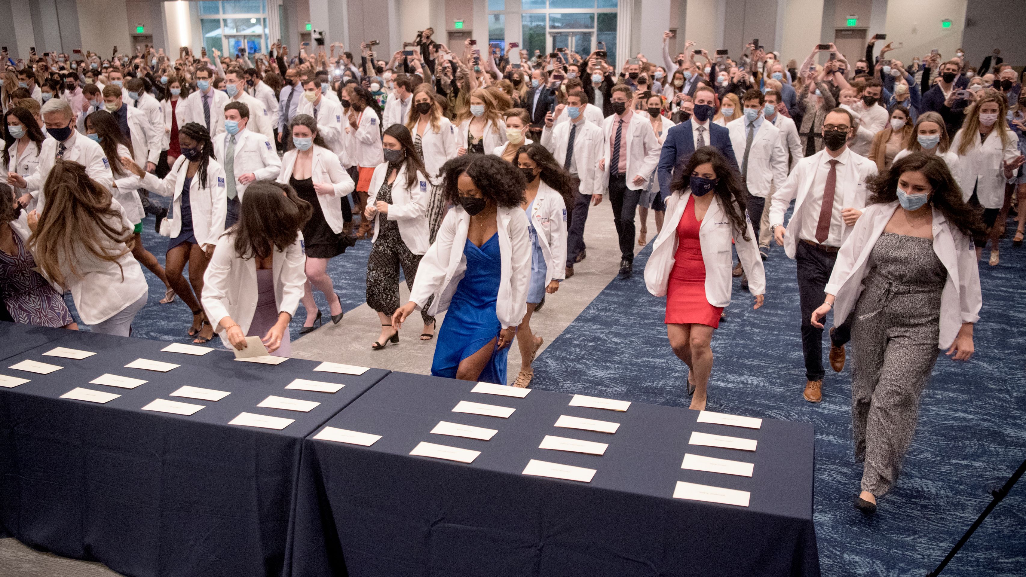Medical students in white coats rush toward envelopes on a Match Day table.