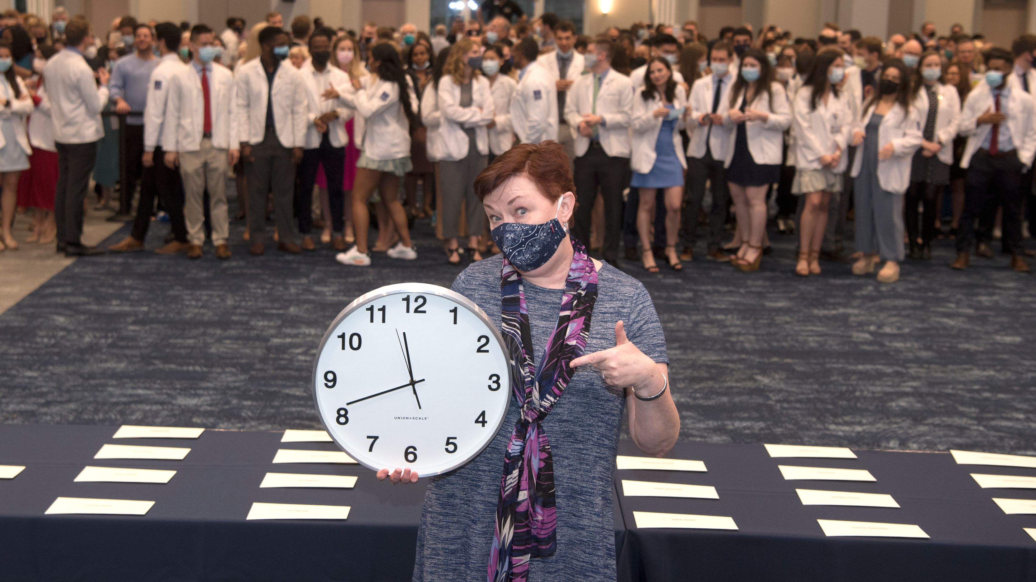 Woman in dress pointing to a clock with medical students in white coats behind her. 