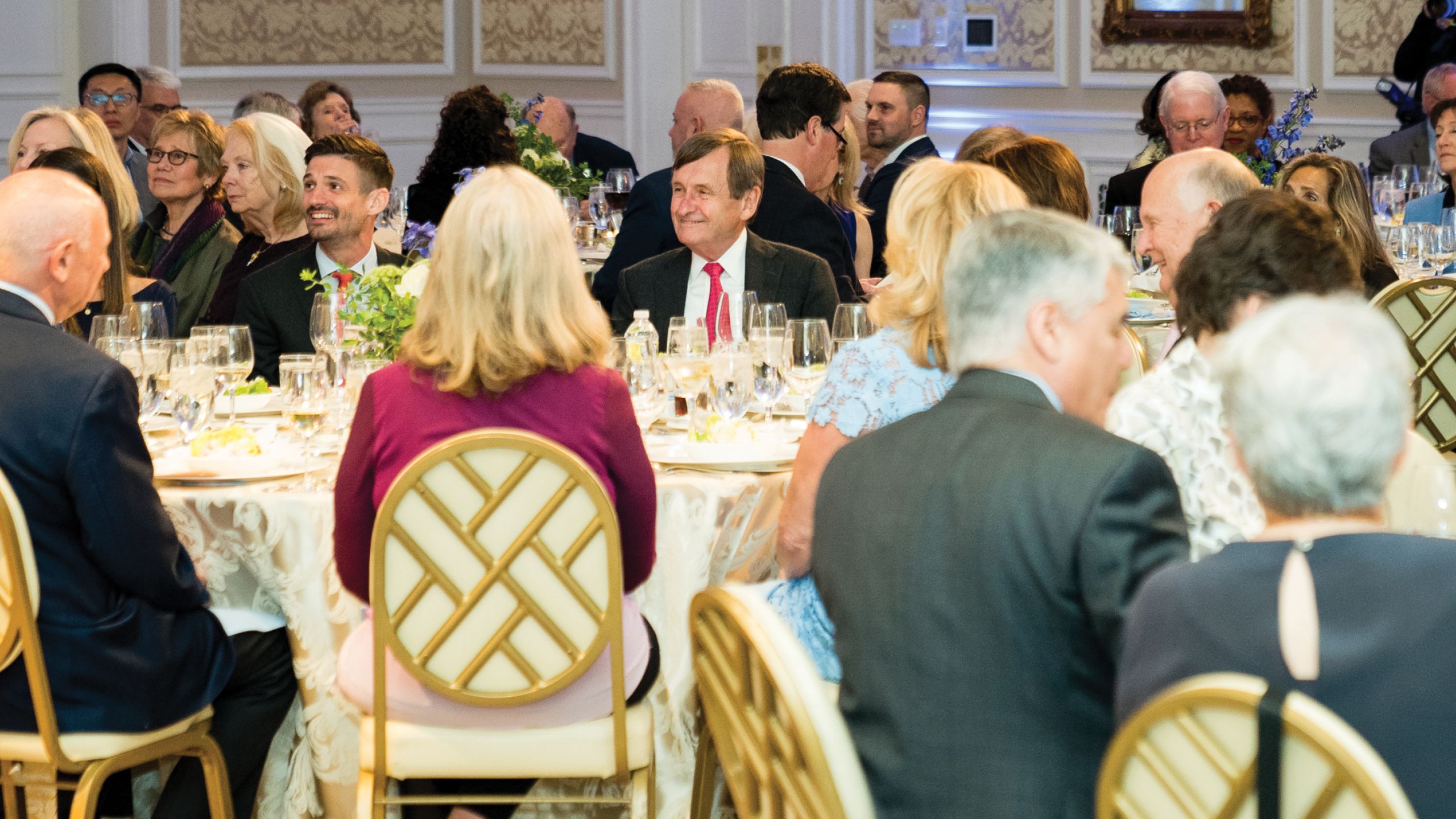 A man smiles broadly seated at a table with many other guests.