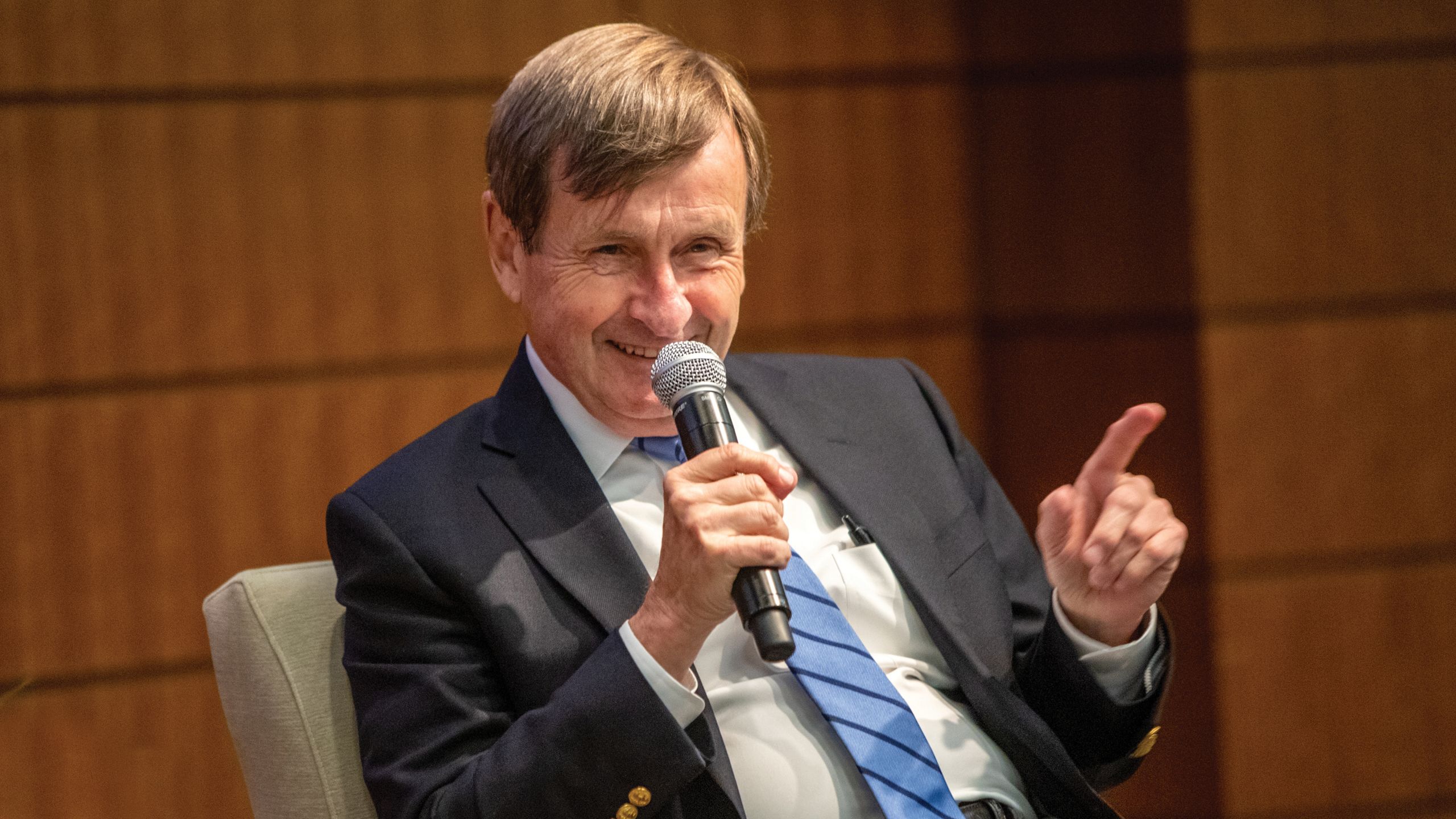 A seated man in a coat and tie talks into a microphone