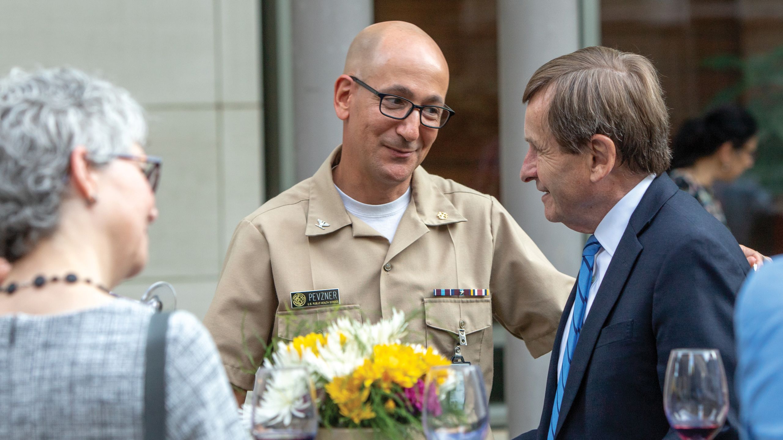 A bald man with glasses in a tan uniform smiles at a man in a coat and tie