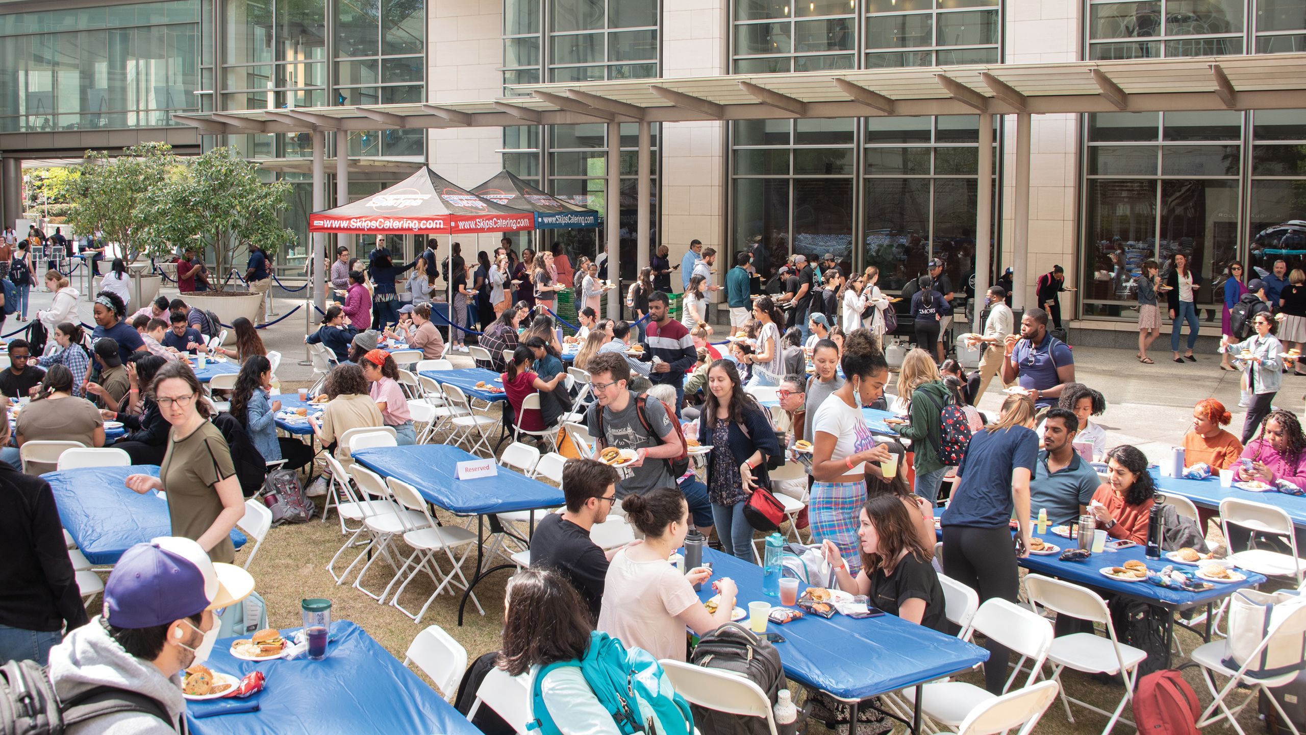 A crowd of people seated outside at long blue tables with white chairs.