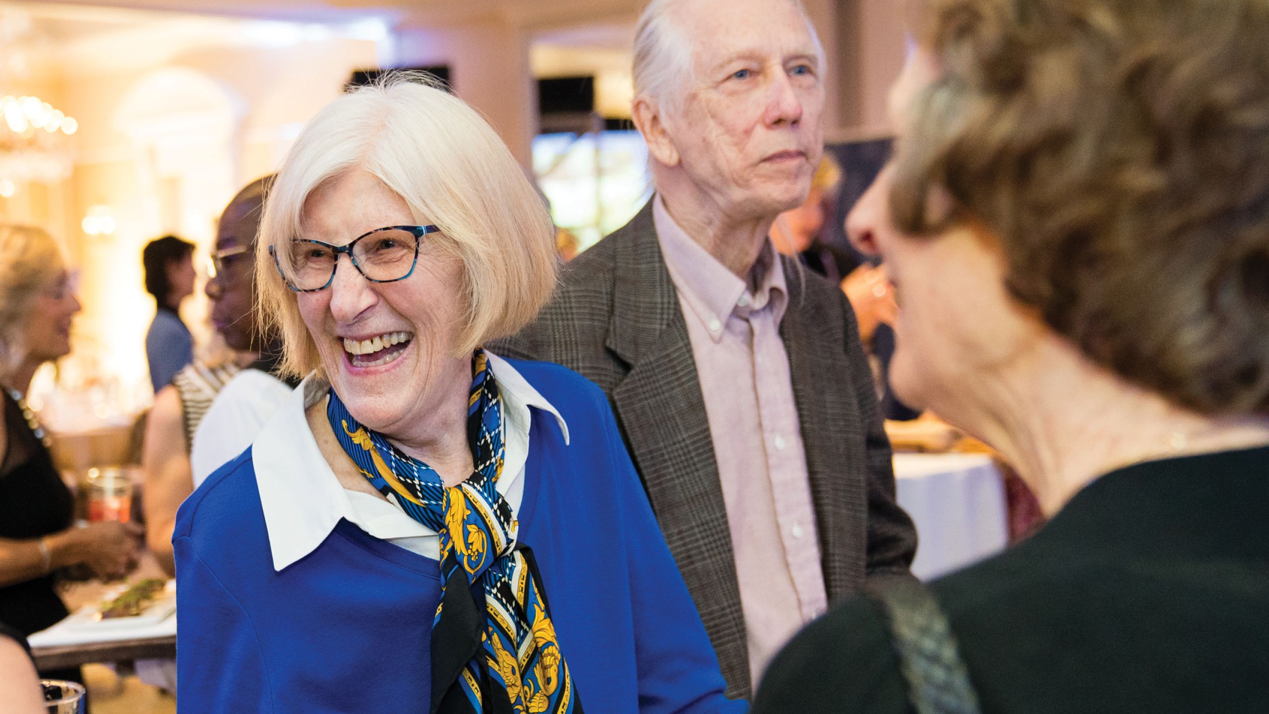 A woman in glasses, light cropped hair and a blue and yellow scarf laughs