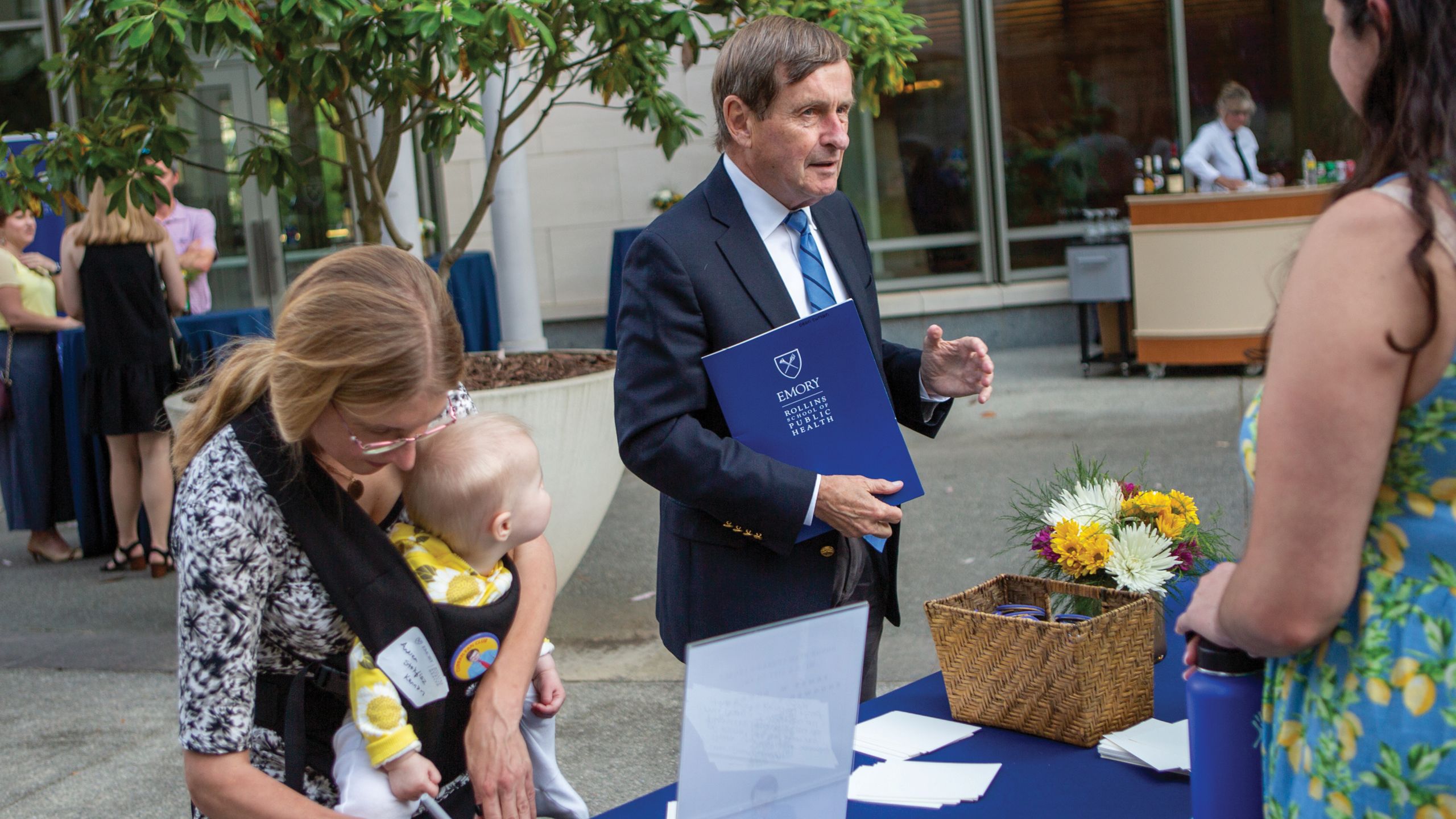 A man in a coat and tie holds a blue folder and talks to other people