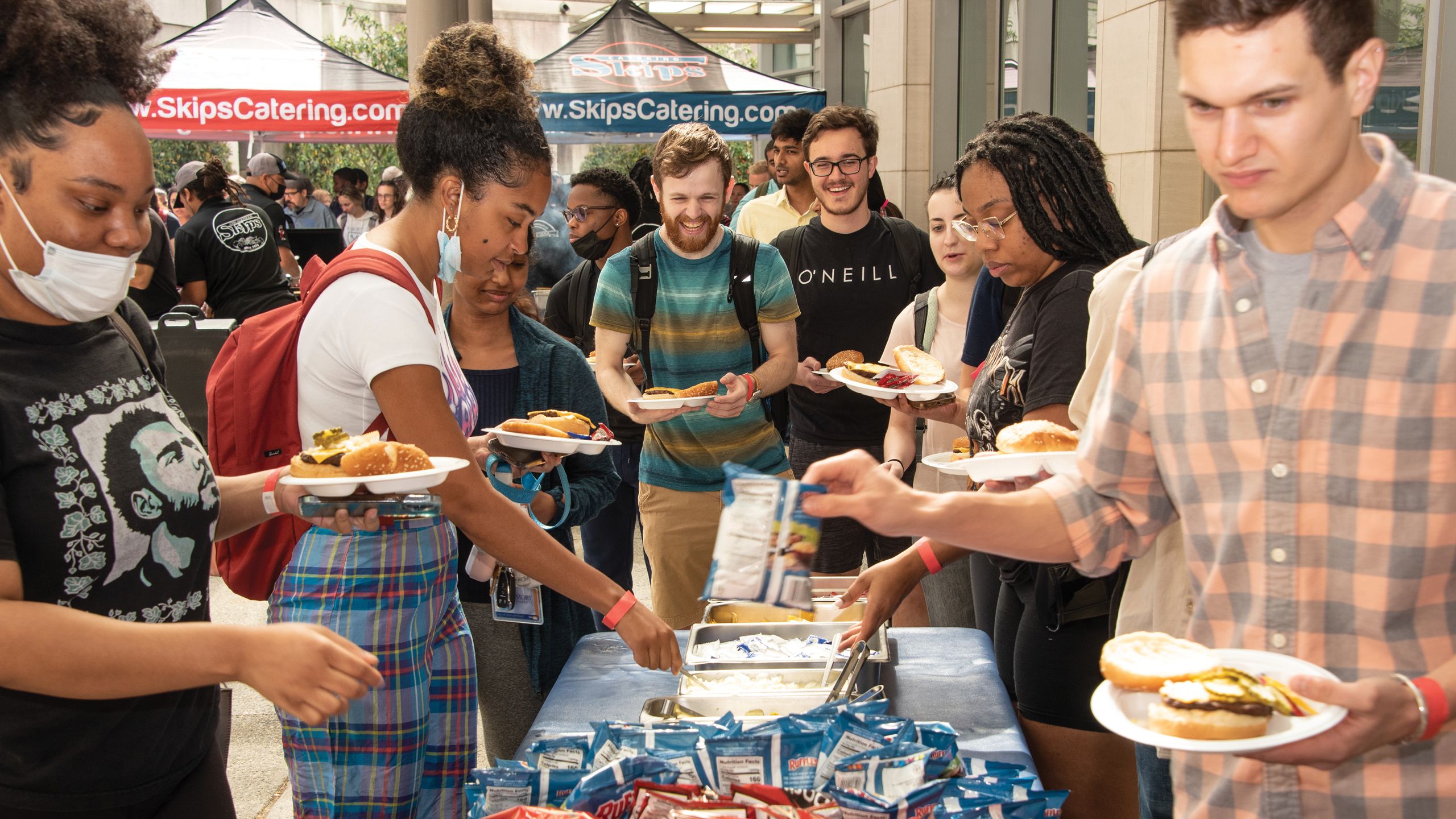People holding plates of hamburgers and hotdogs help themselves to condiments.