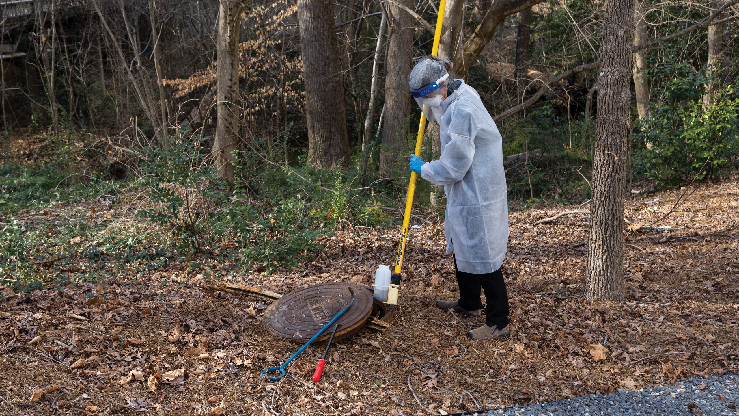 A woman in a mask, plastic visor and robe lowers a pole into an open manhole cover.