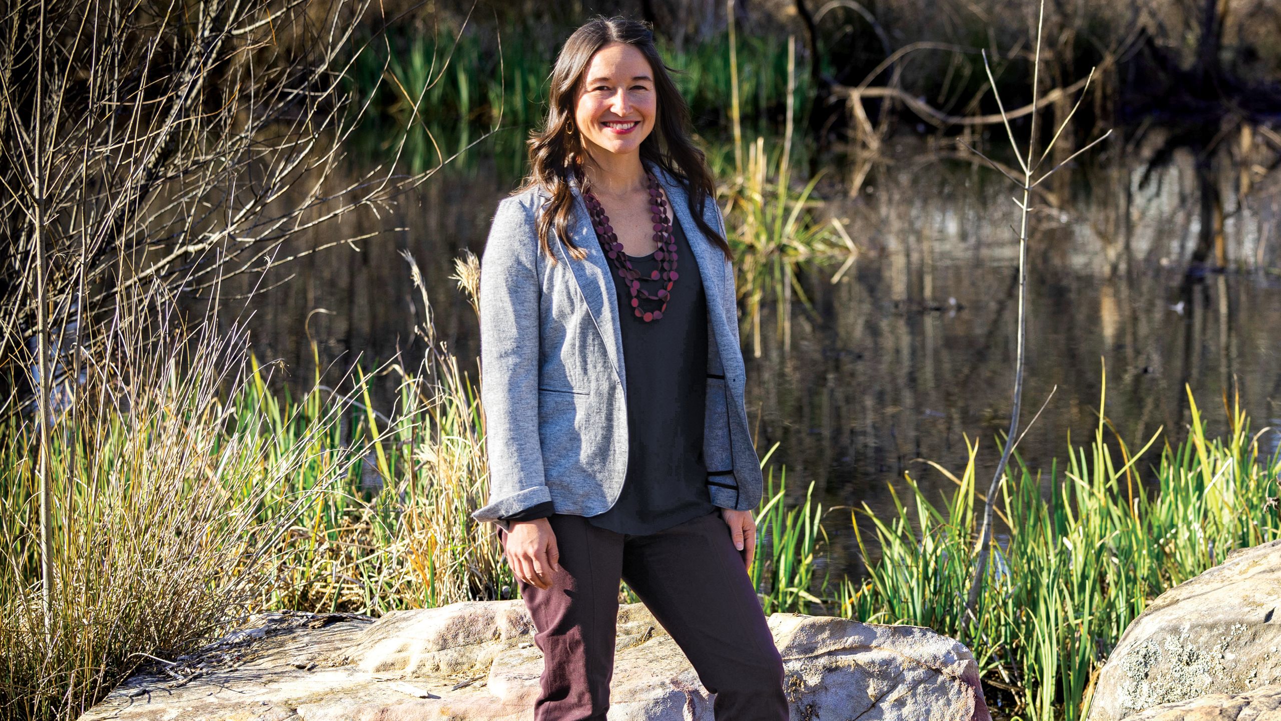 A young woman with long brown hair in a denim jacket stands outside amidst large rocks and tall grass