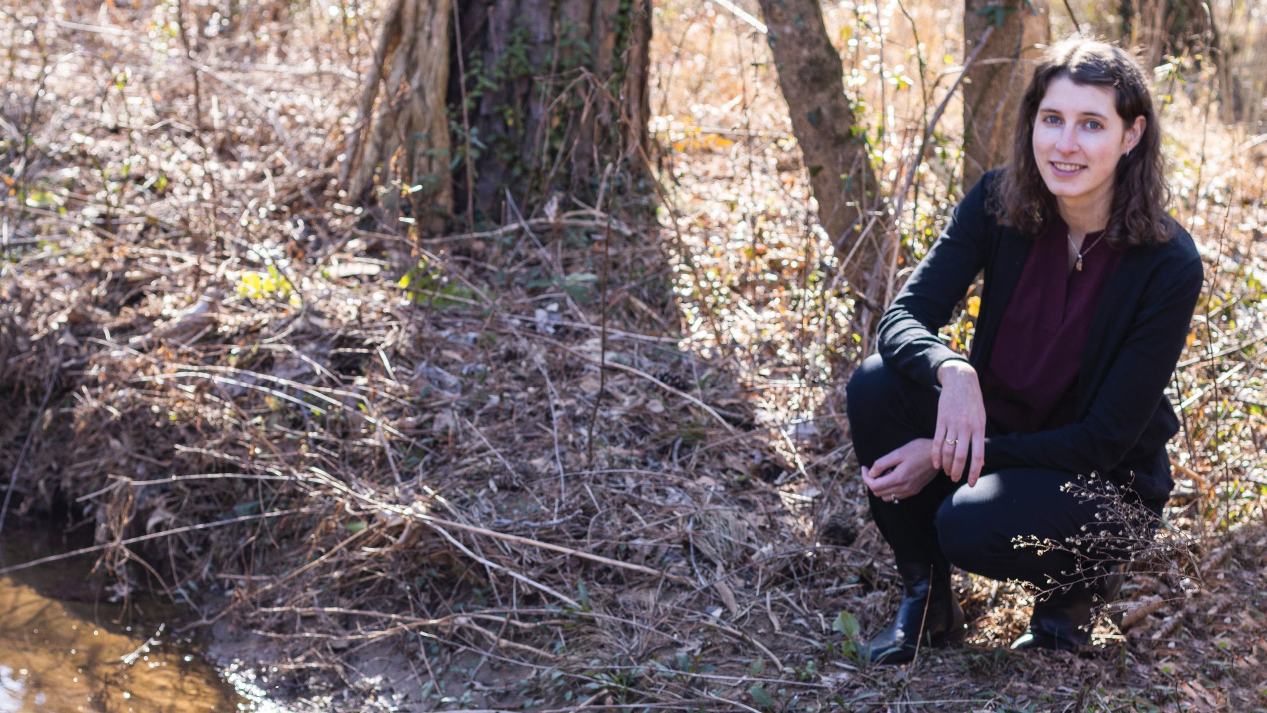A woman in shoulder length brown hair in a dark shirt squats among trees and grasses.