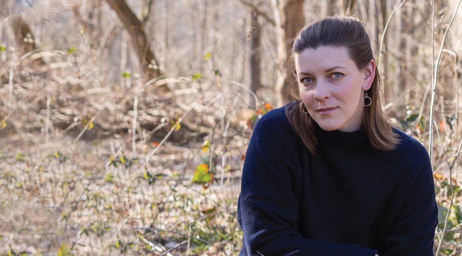 A close up shot of a woman with shoulder length brown hair in a dark blouse outside among grass and trees.