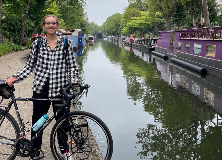 Pomykala holding her bike upright in front of her with a tree-lined canal behind her with busy boat traffic 