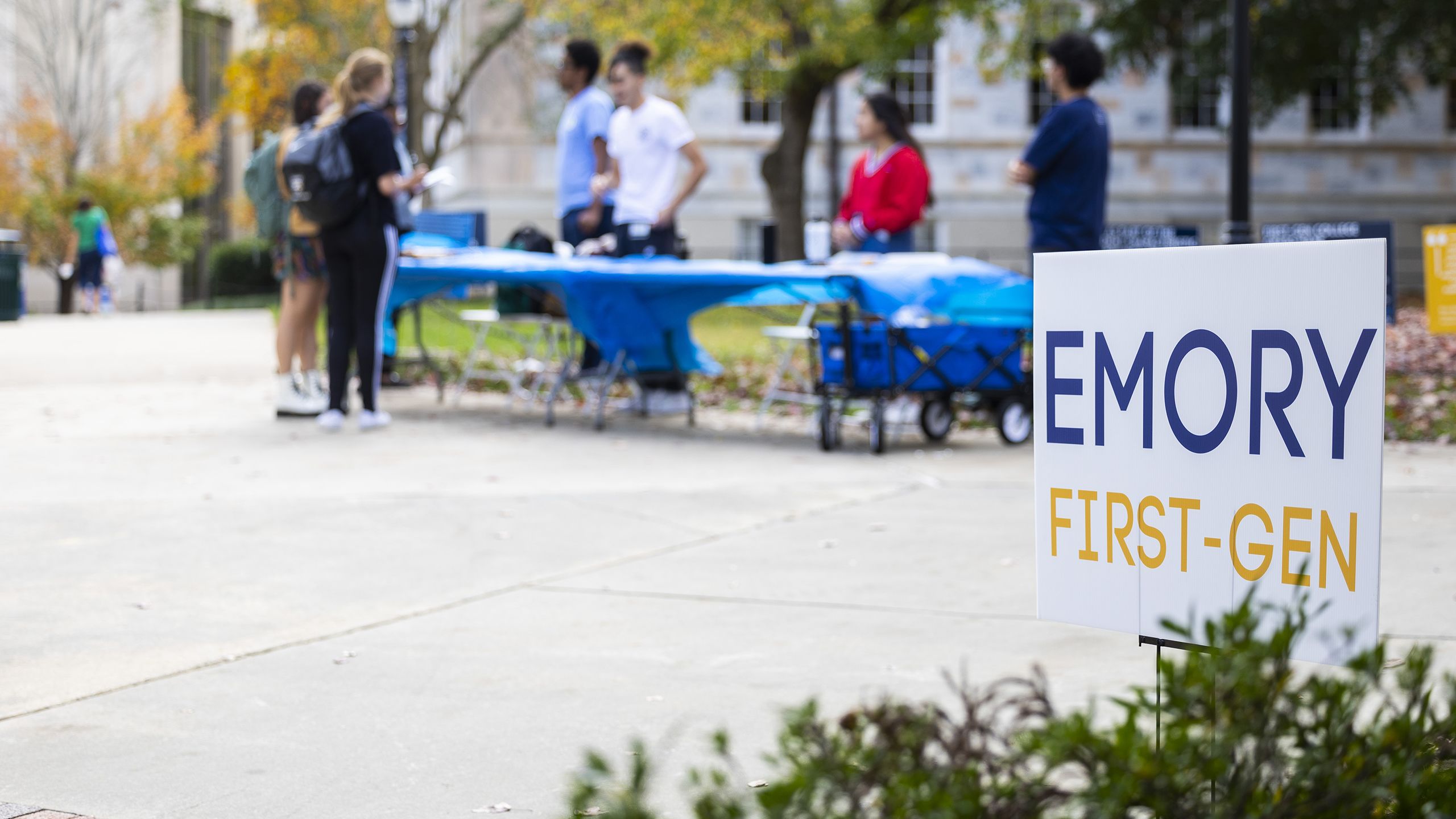 Students stand at a table outdoors near a sign that says Emory First-Gen