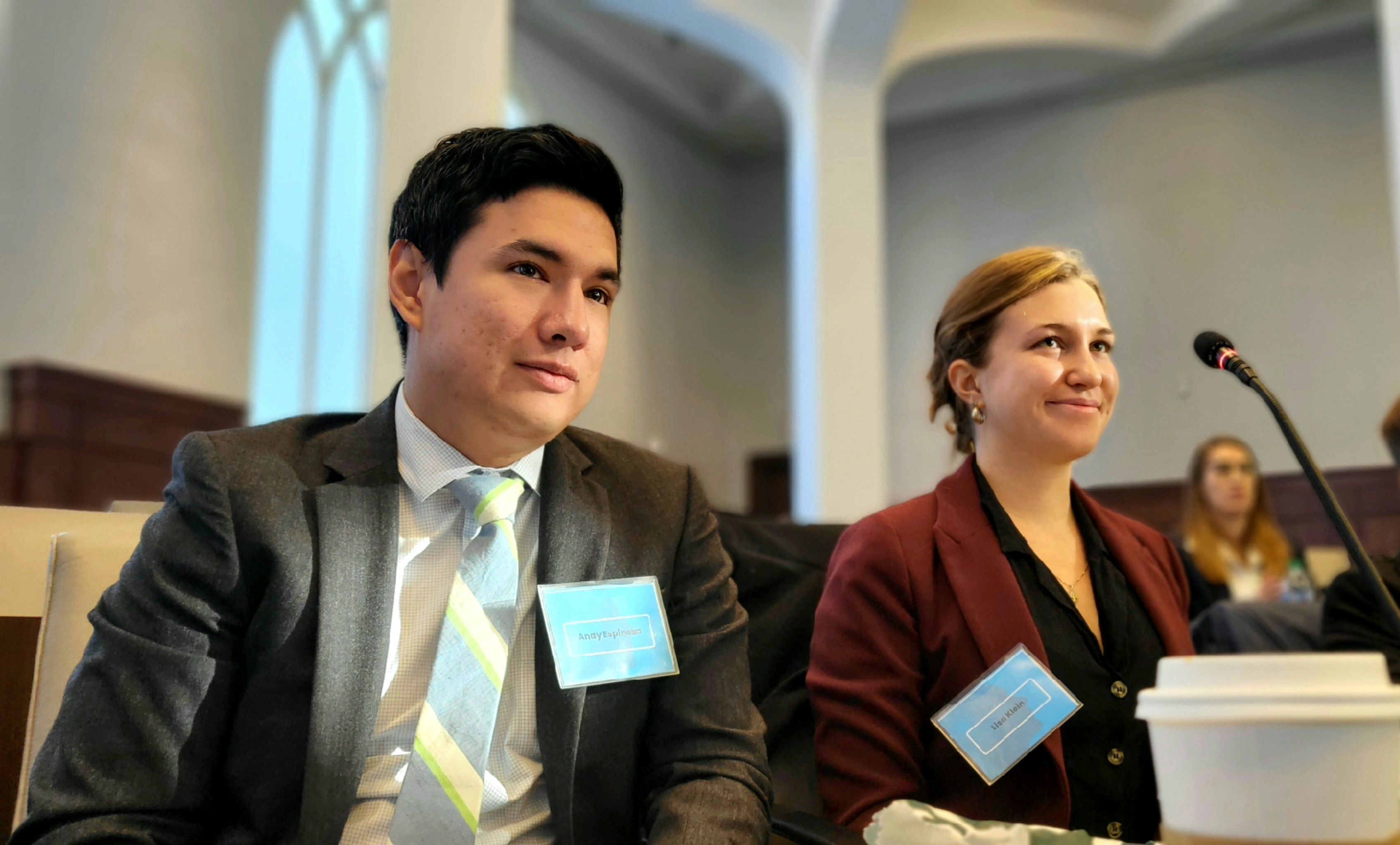 A Latino student and Caucasian female student are seated next to one another in a setting meant to resemble the United Nations.