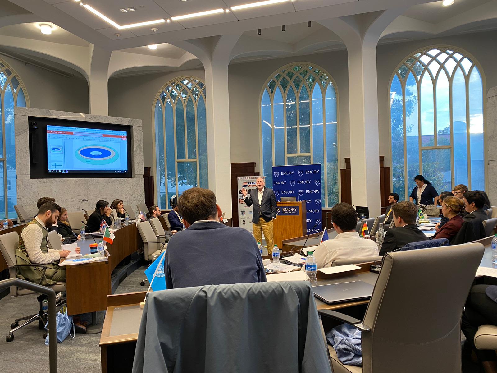 Rows of students are seated on either side of a podium. Large screens on either side of the room are capturing the proceedings. An older Caucasian male professor is addressing them.