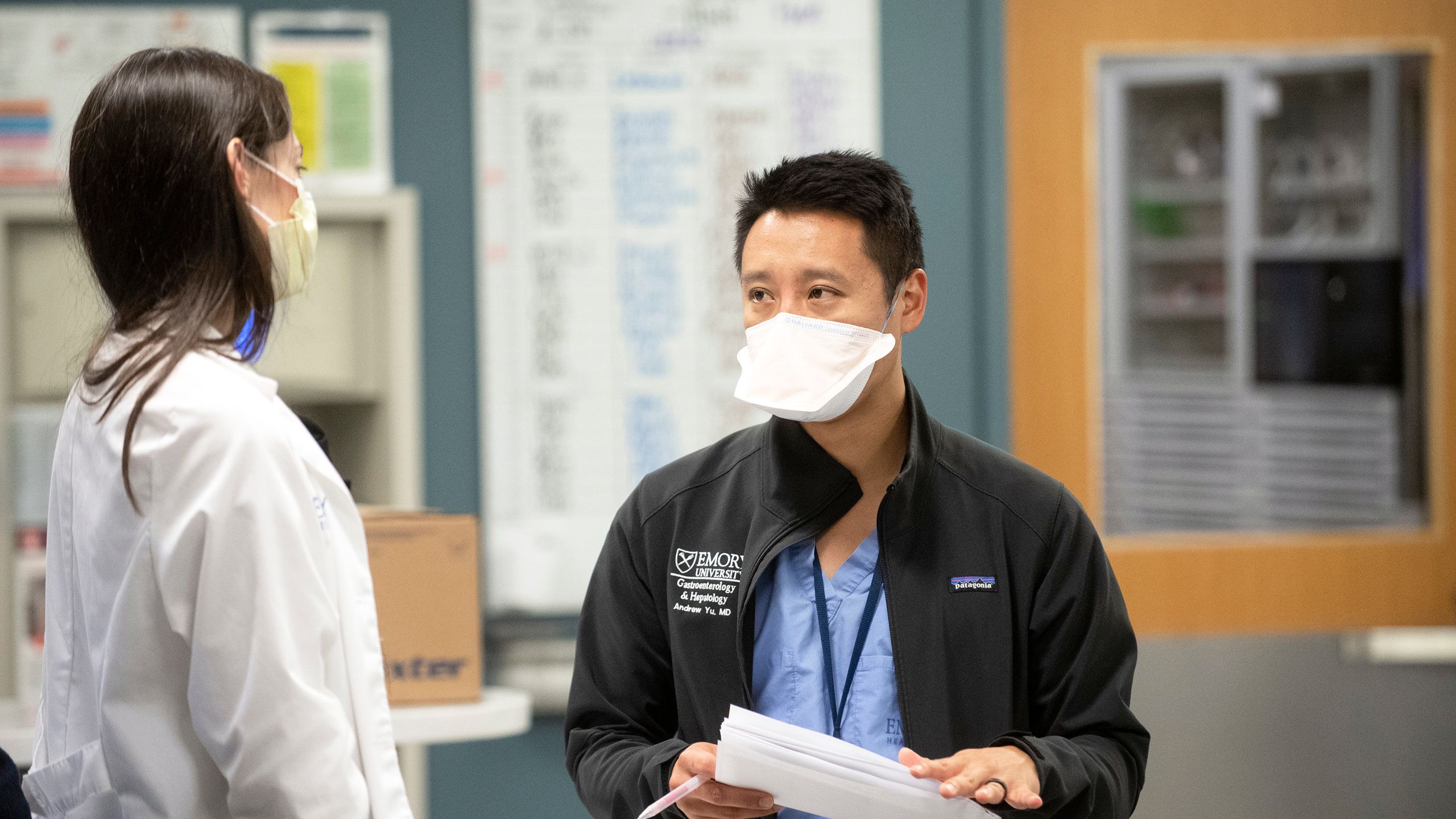 A healthcare worker in a black coat holds papers and talks with a coworker in a critical care unit.