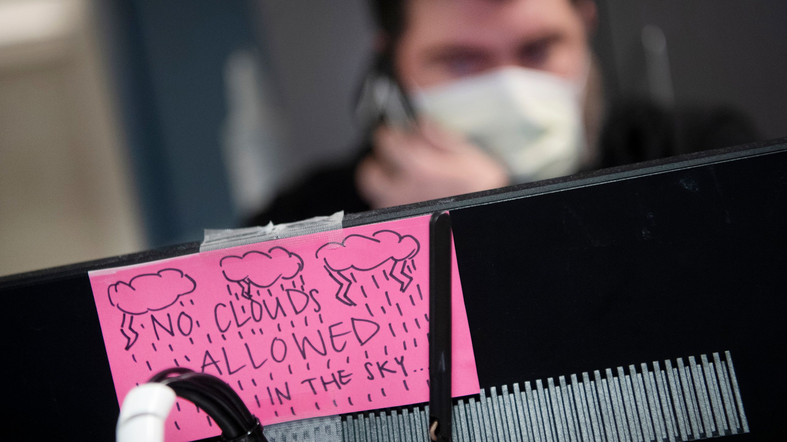 A healthcare worker in a mask sits behind a computer with a pink index card taped to the front reading, "No Clouds Allowed in the Sky..."