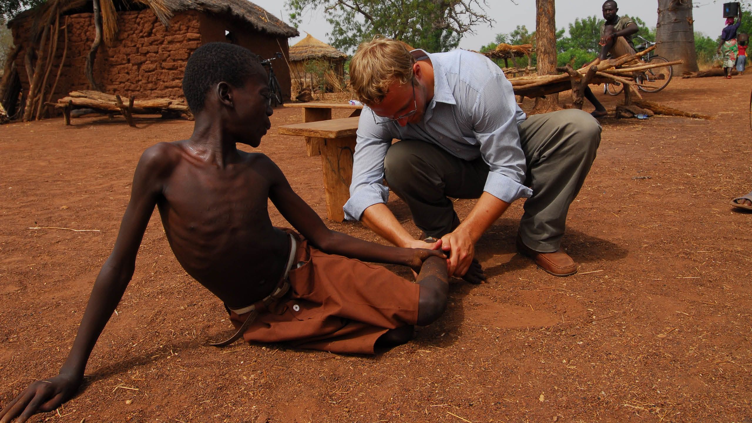  A white man squats to pull a white worm from the foot of a young African boy who is sitting on the ground.