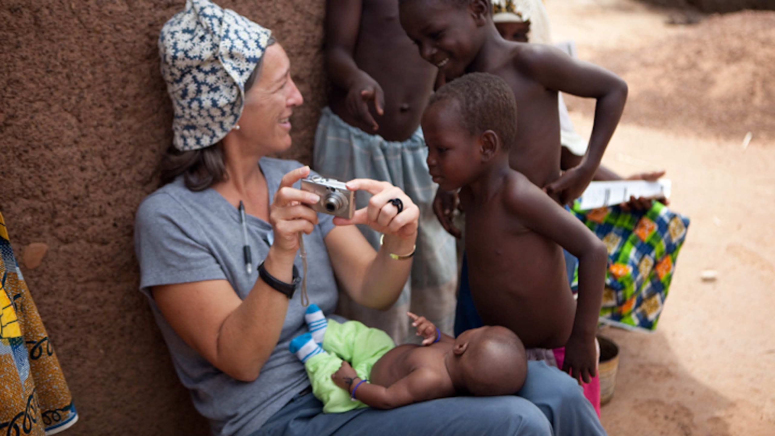 A white woman in a blue and white hat shows her digital camera to a group of curious young African children.