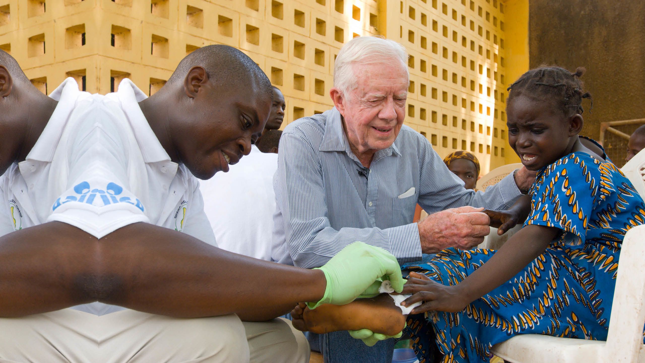 An older man puts his hand on the shoulder of a crying young girl as an unseen person dabs her foot with a clean cloth.