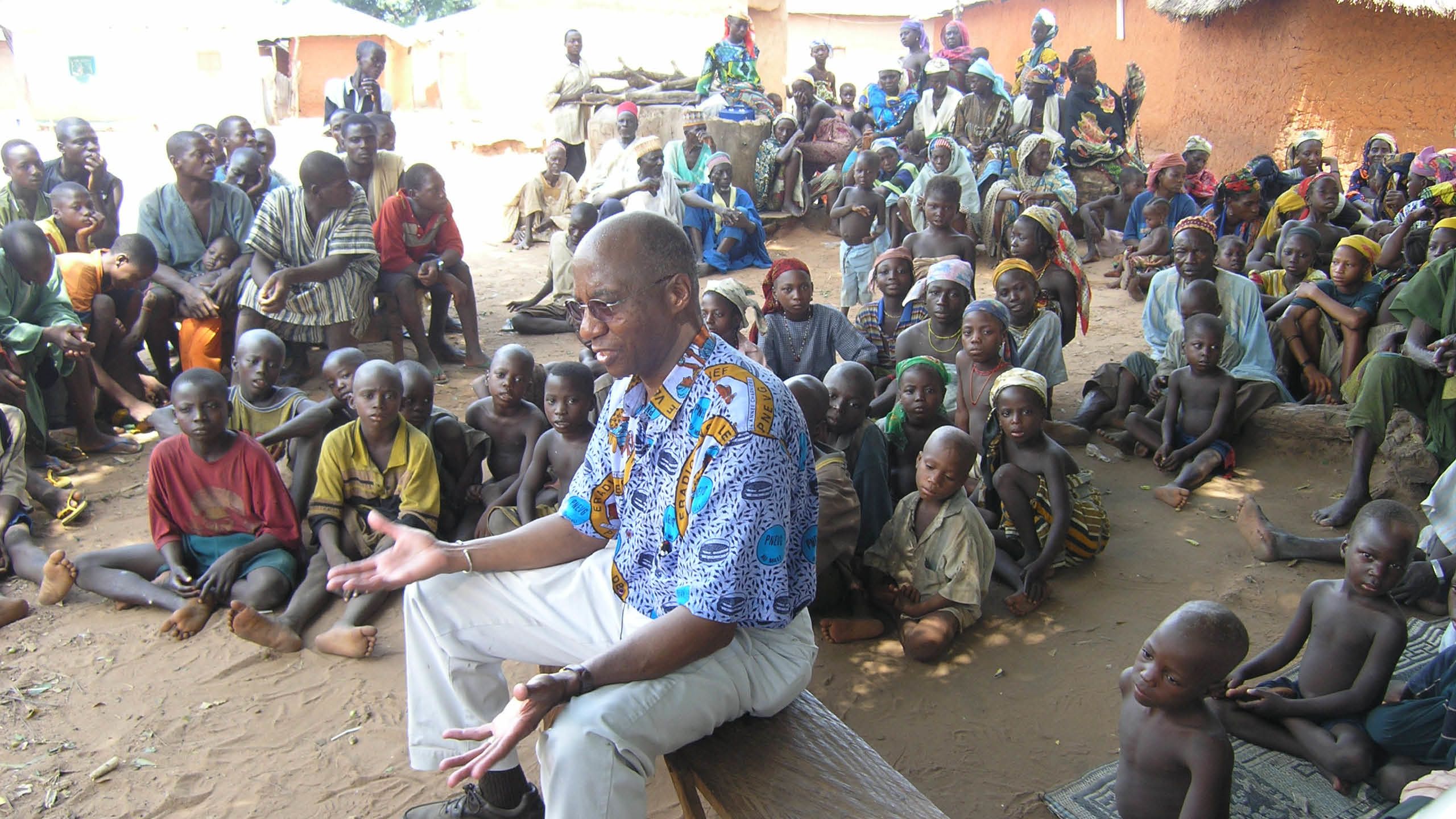 A black man wearing a colorful shirt sits on a bench and talks to a large group of African children sitting on the ground around him.