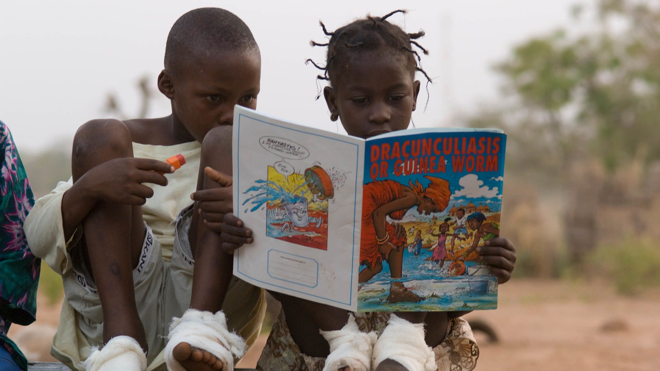 Two African children sit on the ground with bandages wrapped around their feet and look at a colorful comic book.
