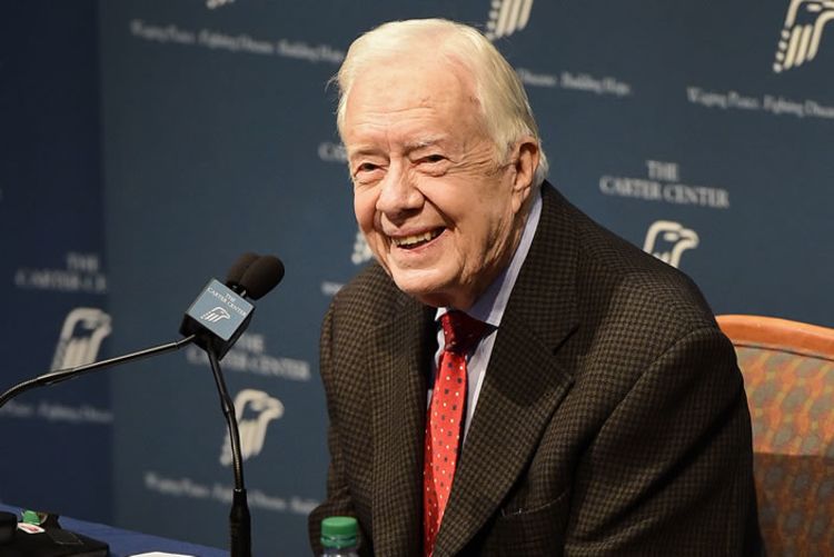 Carter wearing black long sleeve shirt sitting in front of a microphone, with a backdrop reading The Carter Center