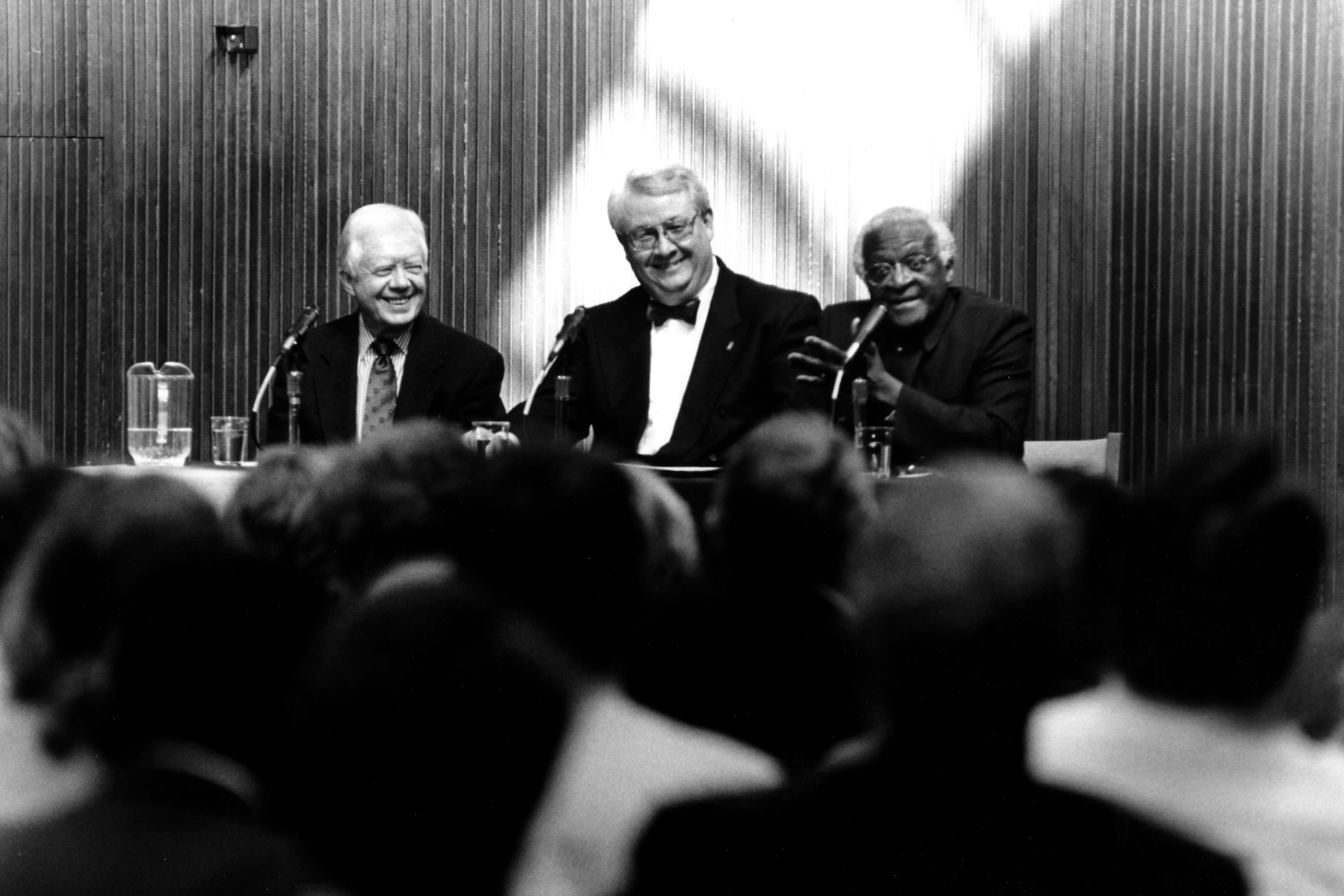 Black and white image of the three men at a table, with microphones, speaking to a crowded room.