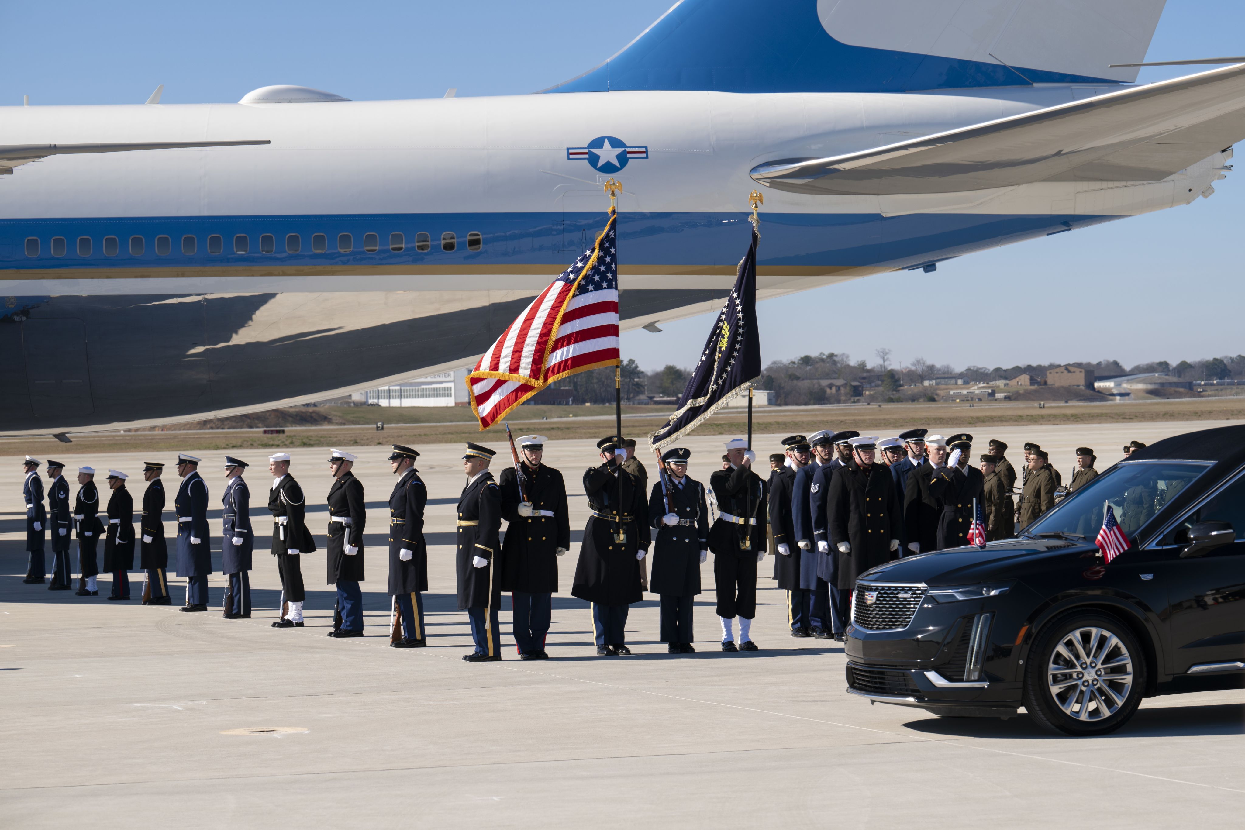 A military honor guard stands at attention in front of a plane with a black hearse in the foreground