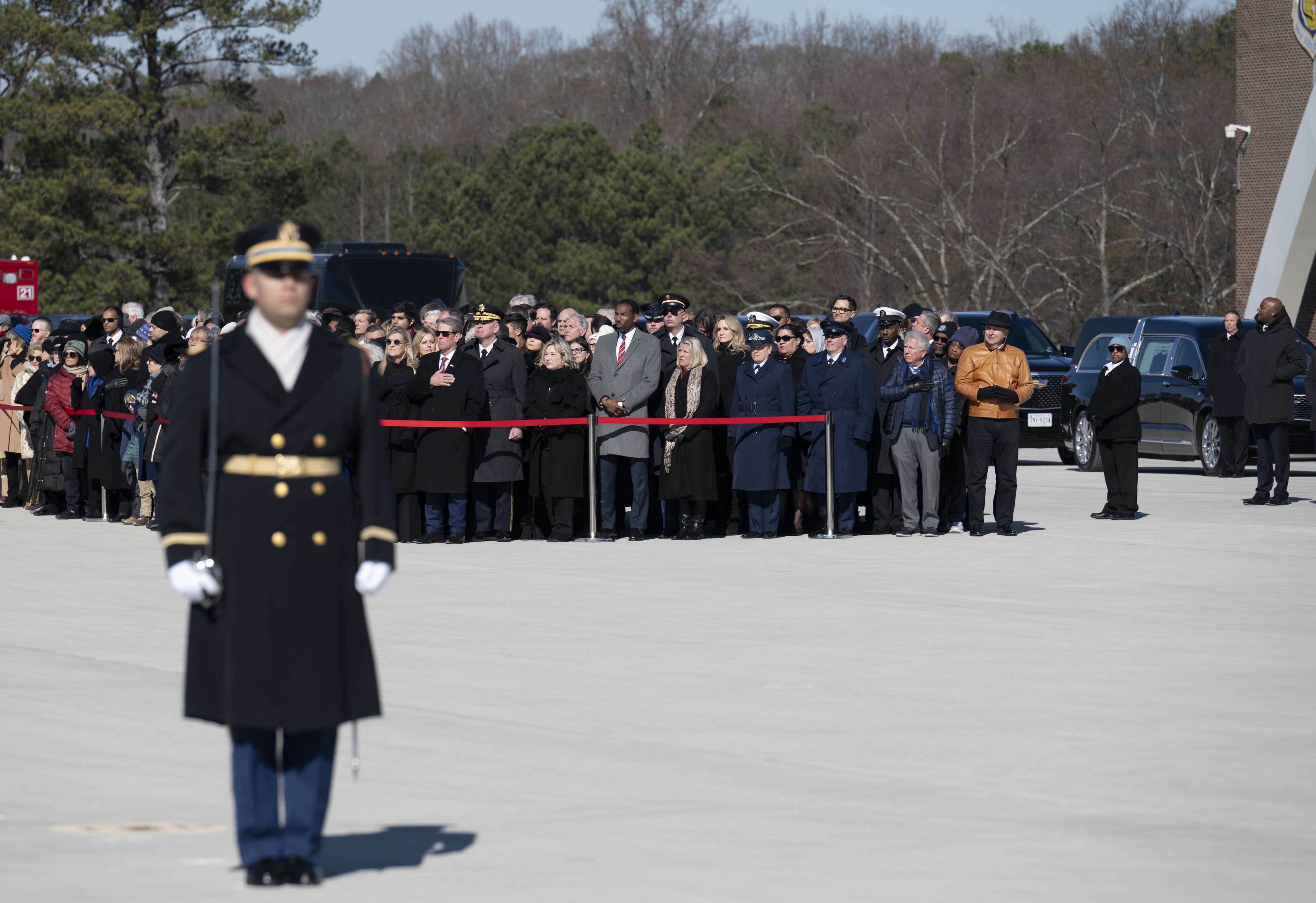 A crowd stands behind a line as a military color guard member stands in the foreground