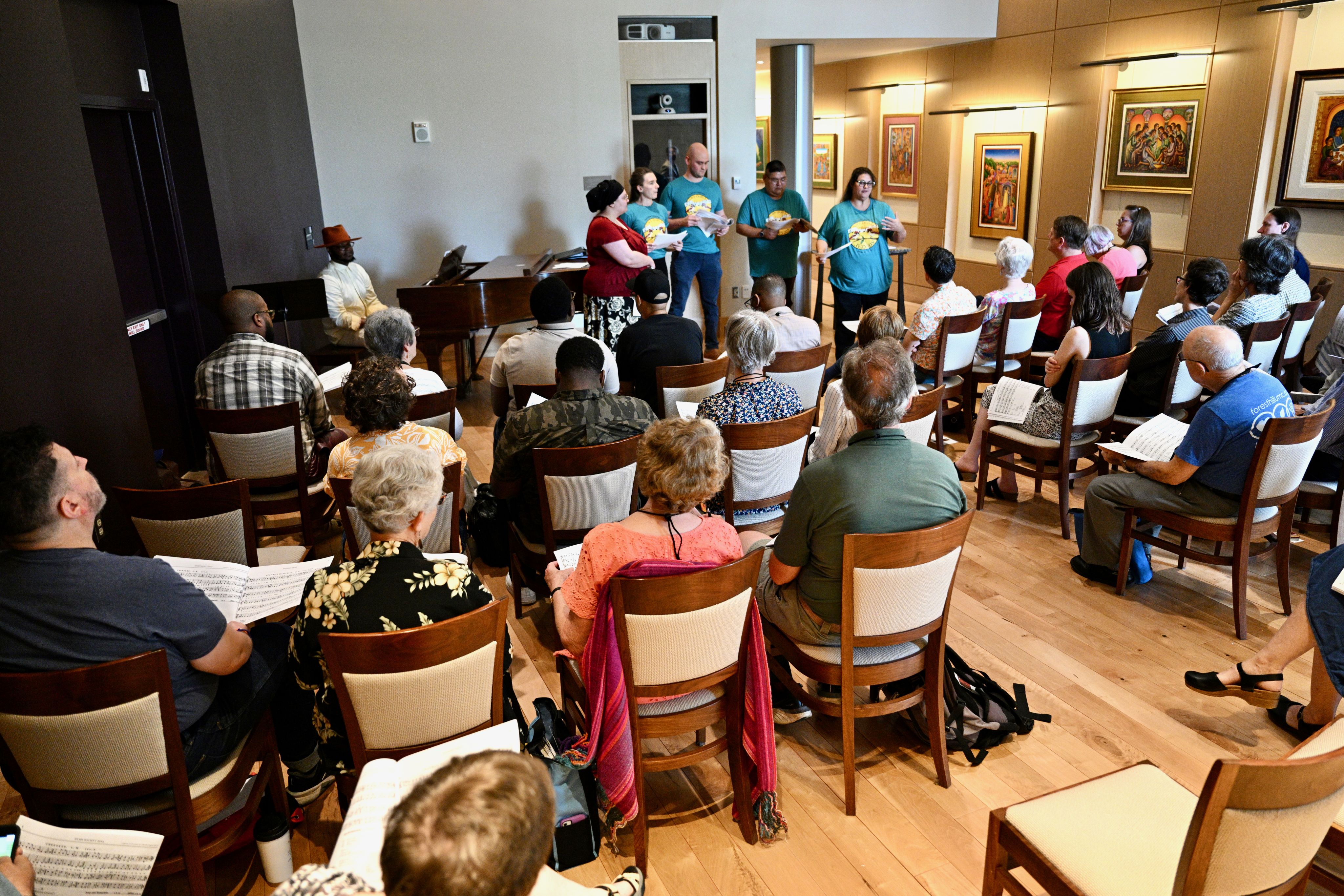 Five people stand, singing, in front of a seated audience.