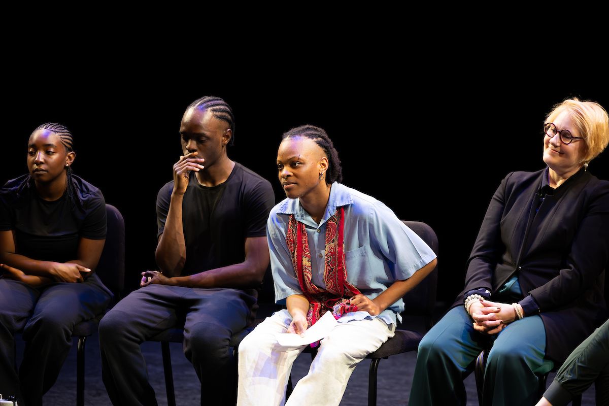 Four seated figures face forward, three African-American college students and one older caucasian teacher. The student second from the right leans forward, listening to someone talk.