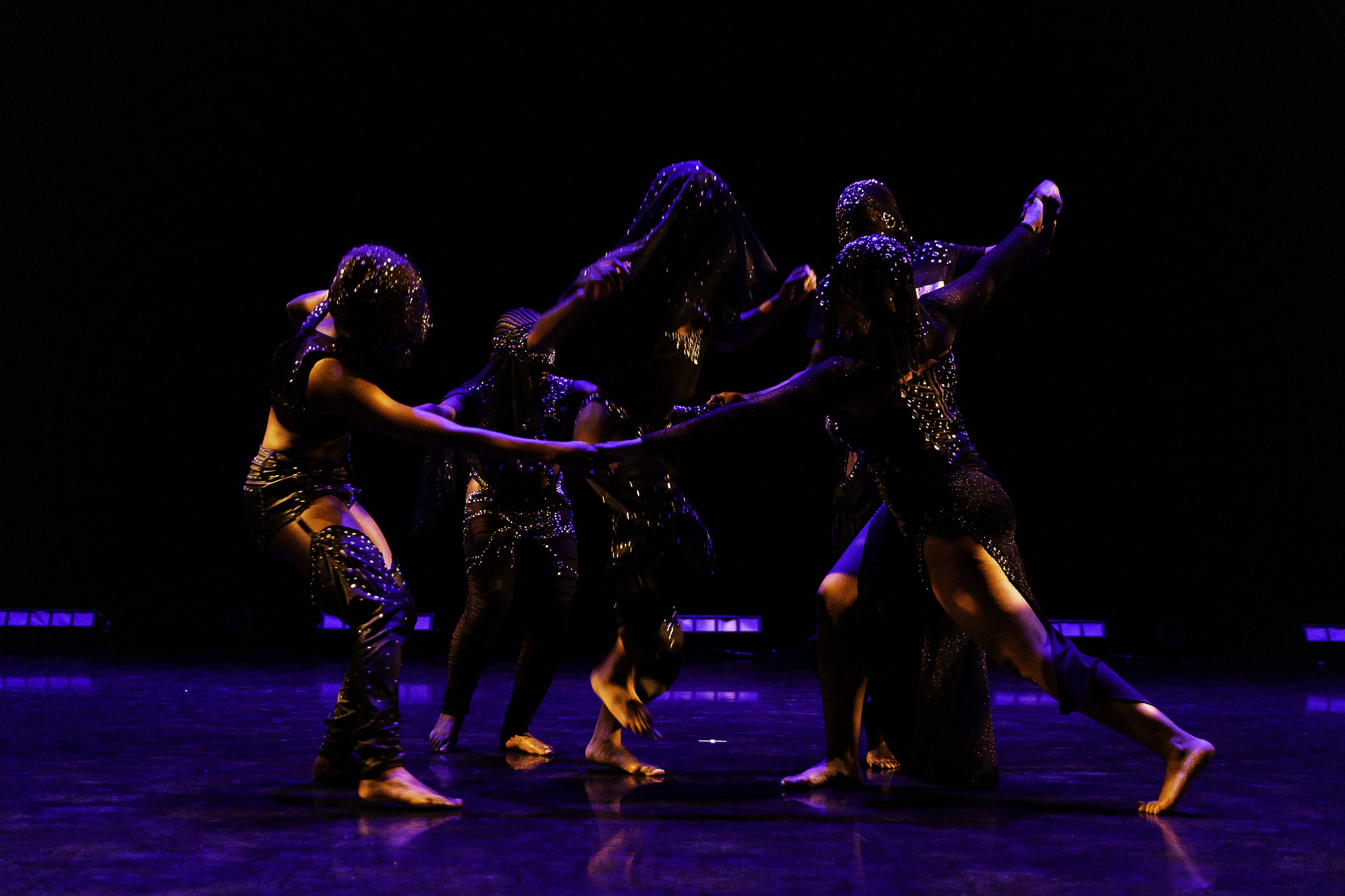 Four dancers grasp hands in a circle. A fifth dancer crouches in the middle.