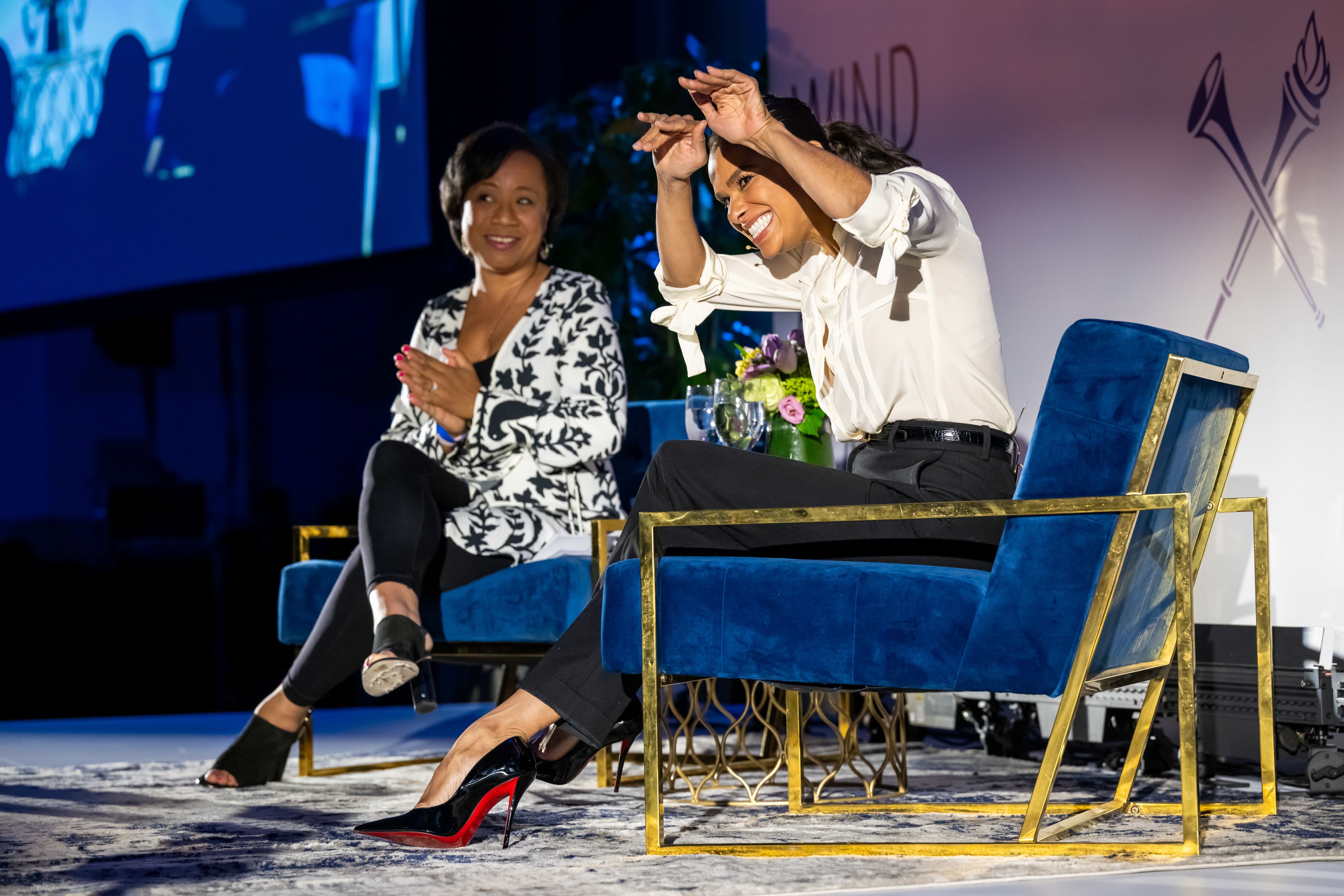 Two women sit in chairs onstage. One holds her hands up in the air, waving to the crowd and grinning.