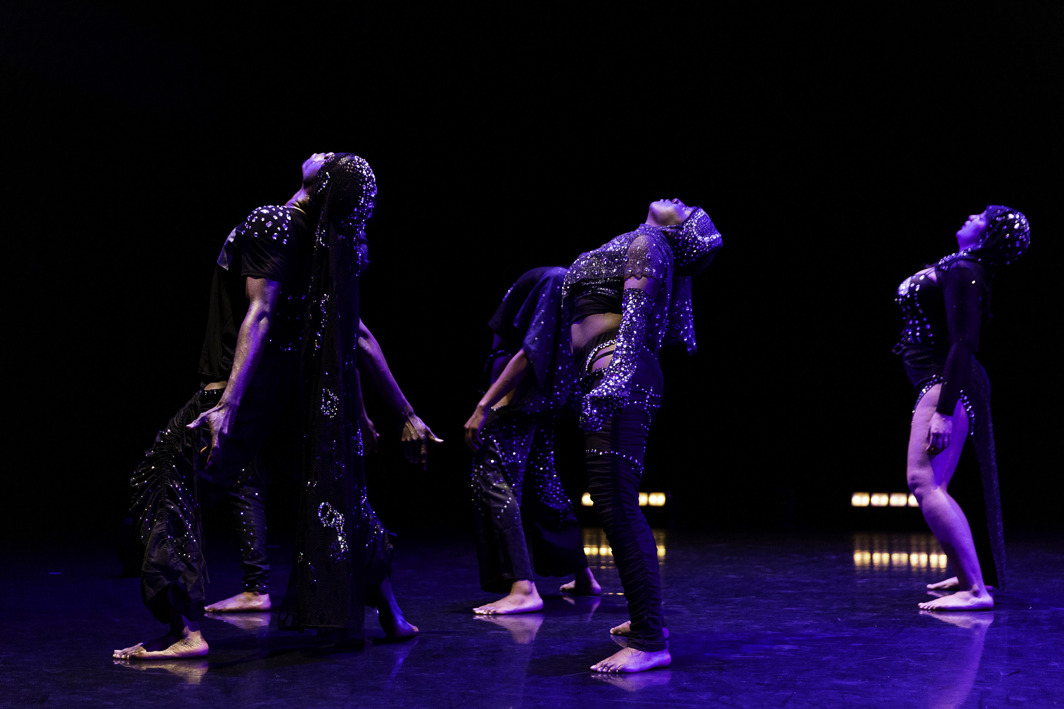Five African-American dancers, wearing sparkly black costumes, stand facing left with their backs arched in unison.