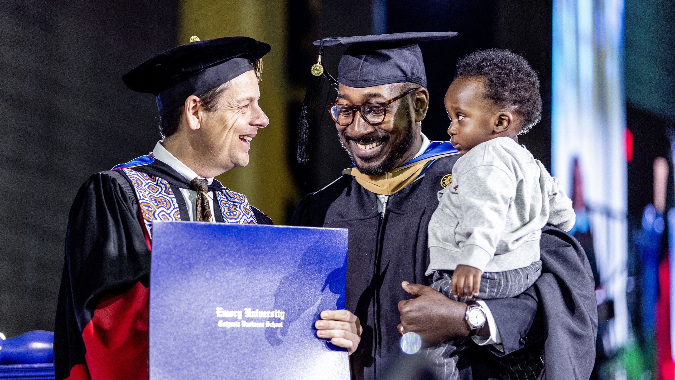 A graduate carries his infant son as he receives his diploma