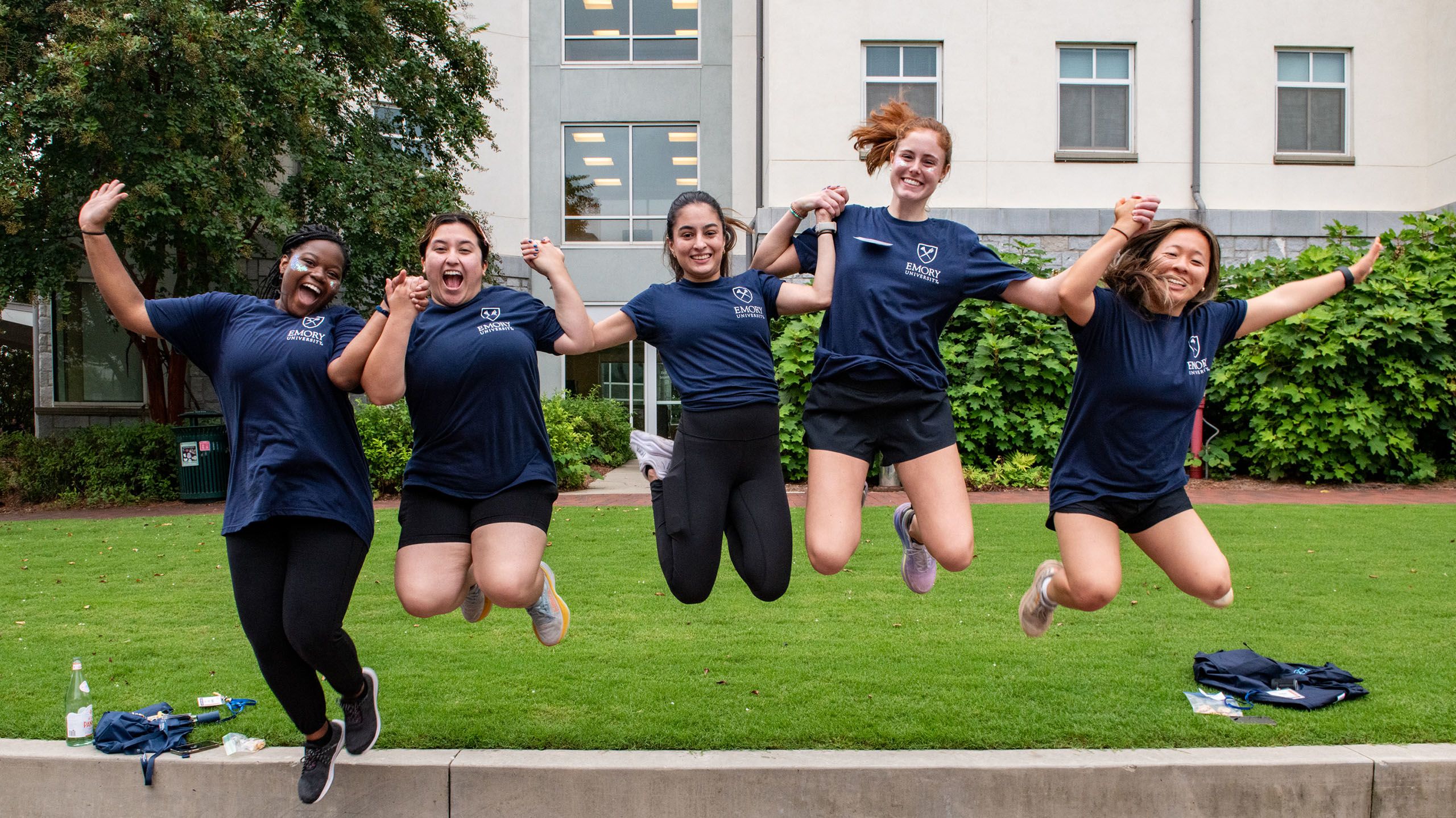 A group of female students in matching Emory shirts hold hands and jump