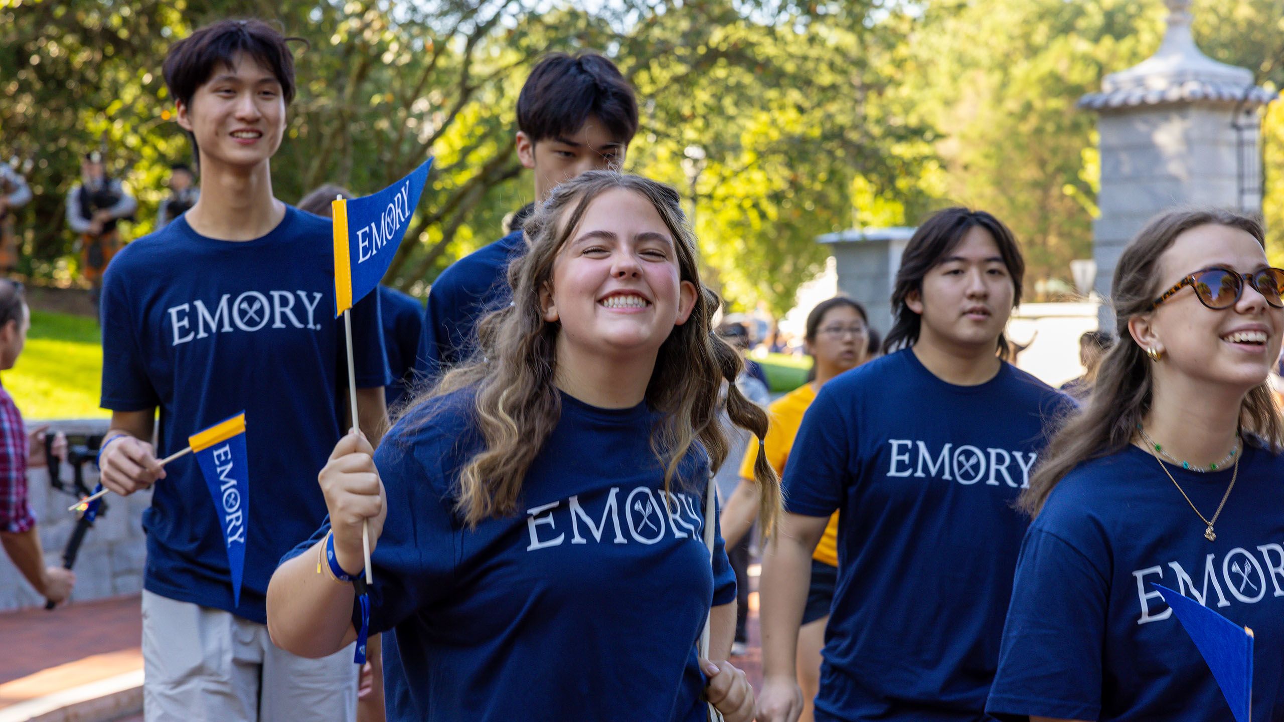 Group of students walking through the Emory gate, wearing blue Emory tshirts. Female student in center is smiling and waving an Emory pennant.
