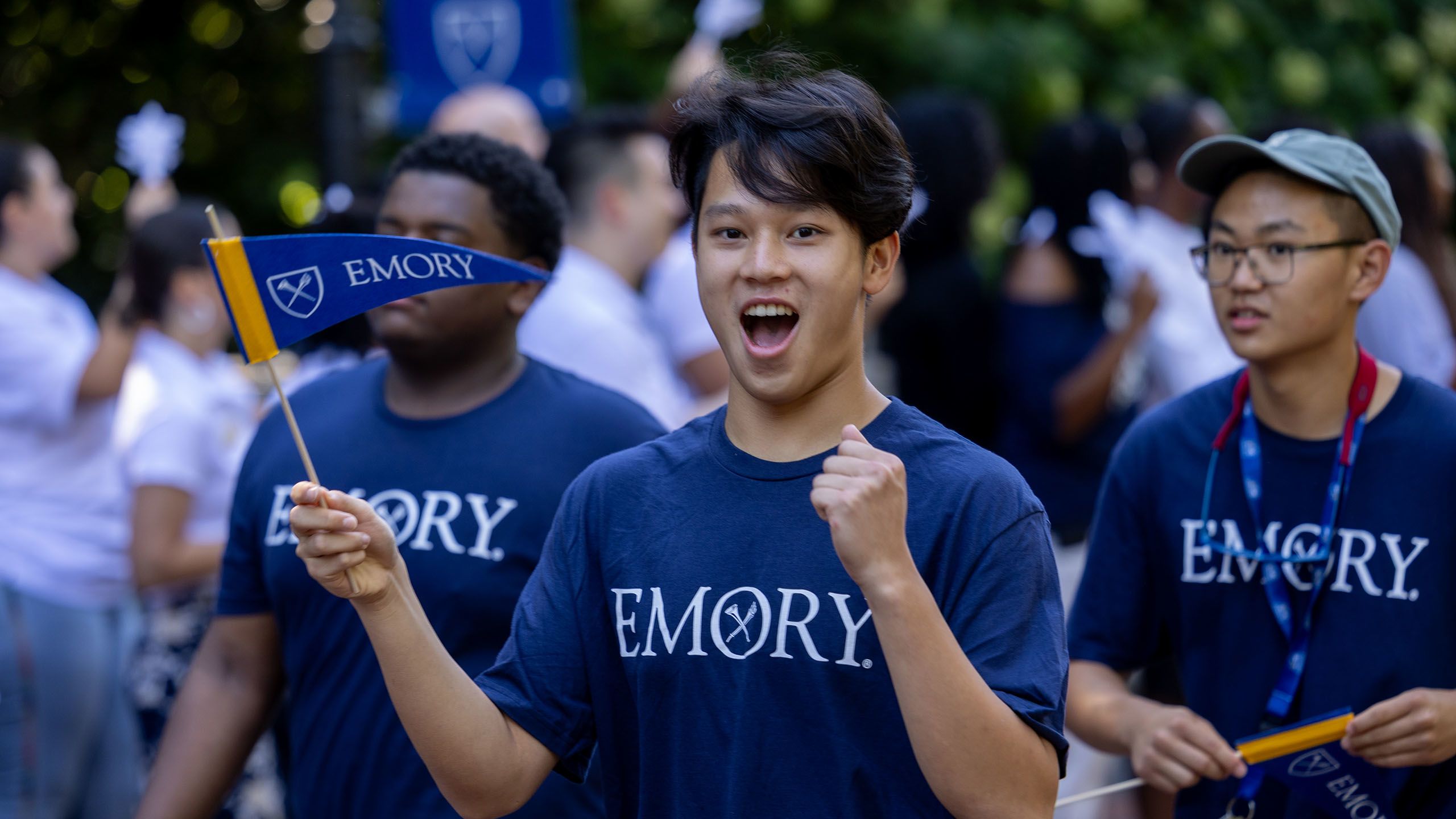 Male student wearing a blue Emory shirt cheers and waves an Emory pennant.
