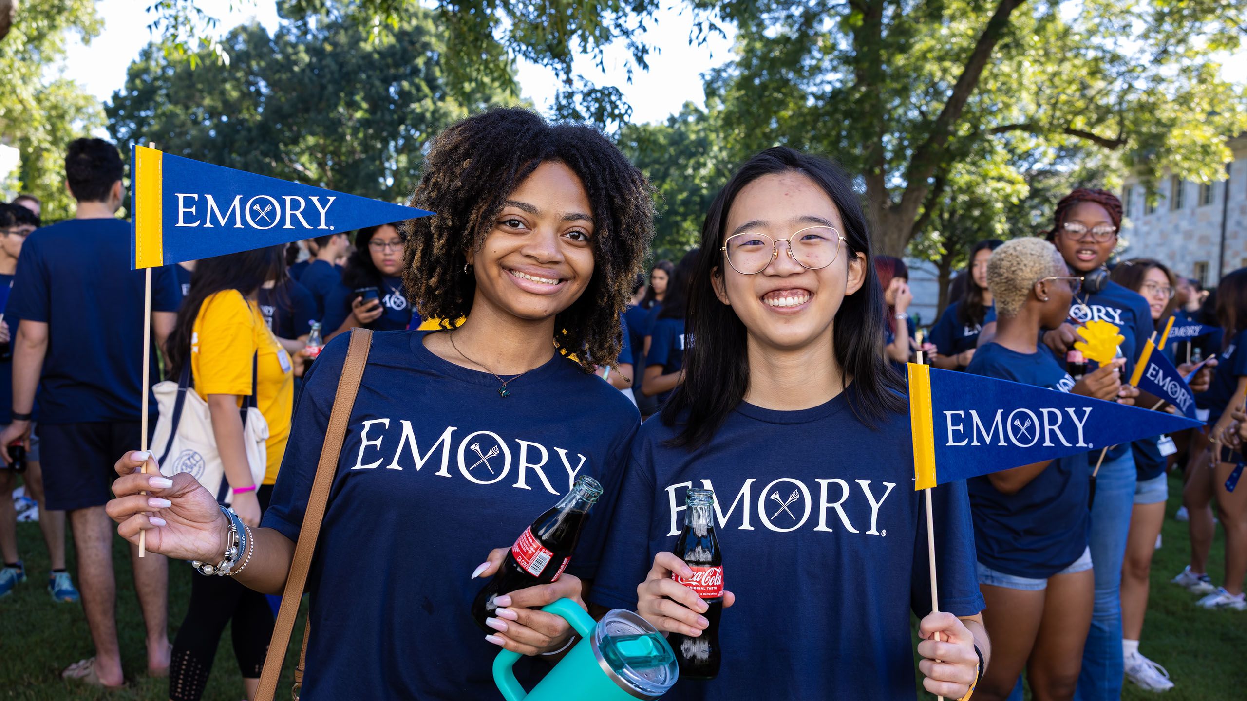 Two female students hold bottles of Coca Cola and wave Emory pennants.