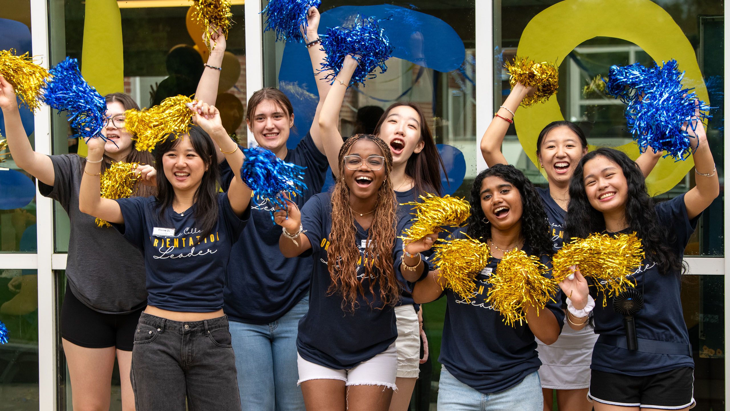 Large group orientation leaders, wearing matching shirts, wave blue and gold pom poms and cheer.