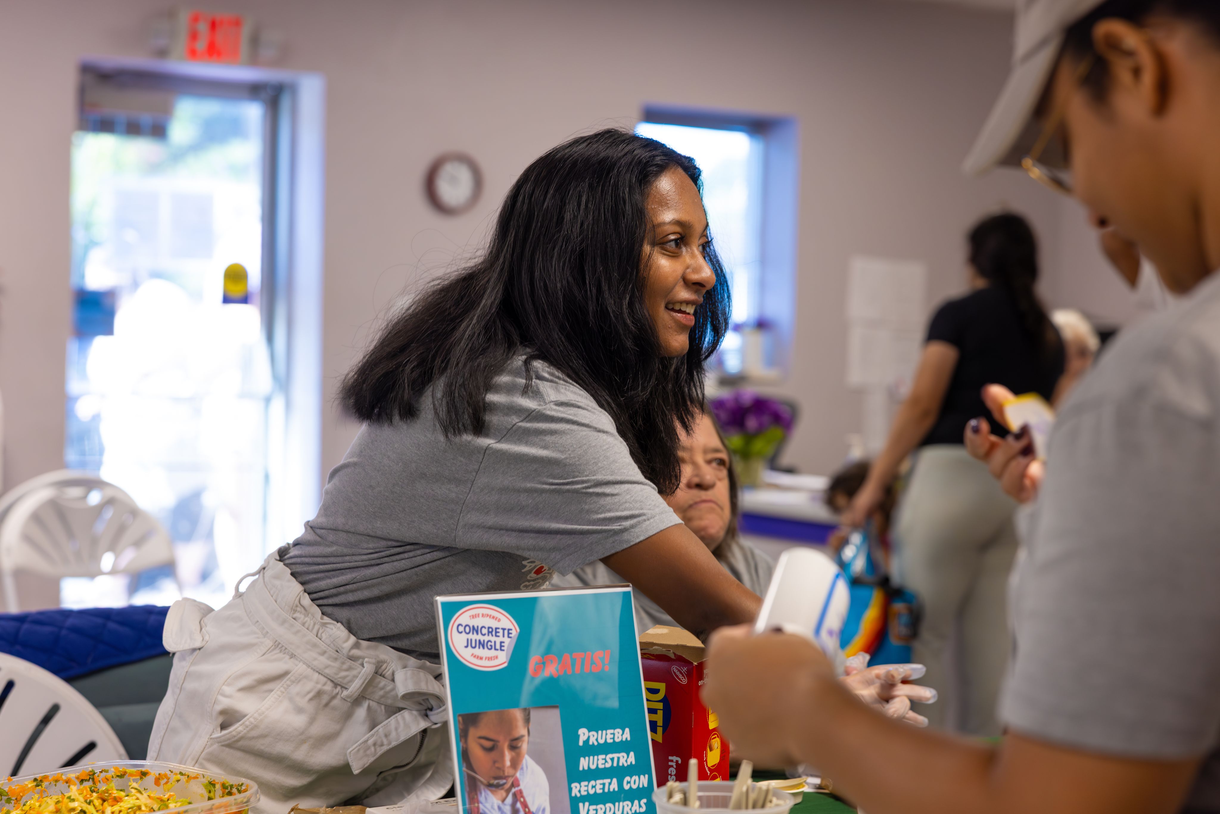 A young woman standing at a table reaches out, smiling, to someone out of shot.