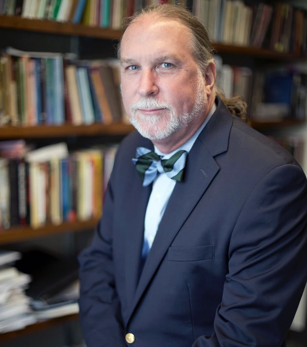 A man with a gray mustache and beard smiles at the camera. His long hair is pulled back and he wears a dark blue suit and a light blue bow tie.