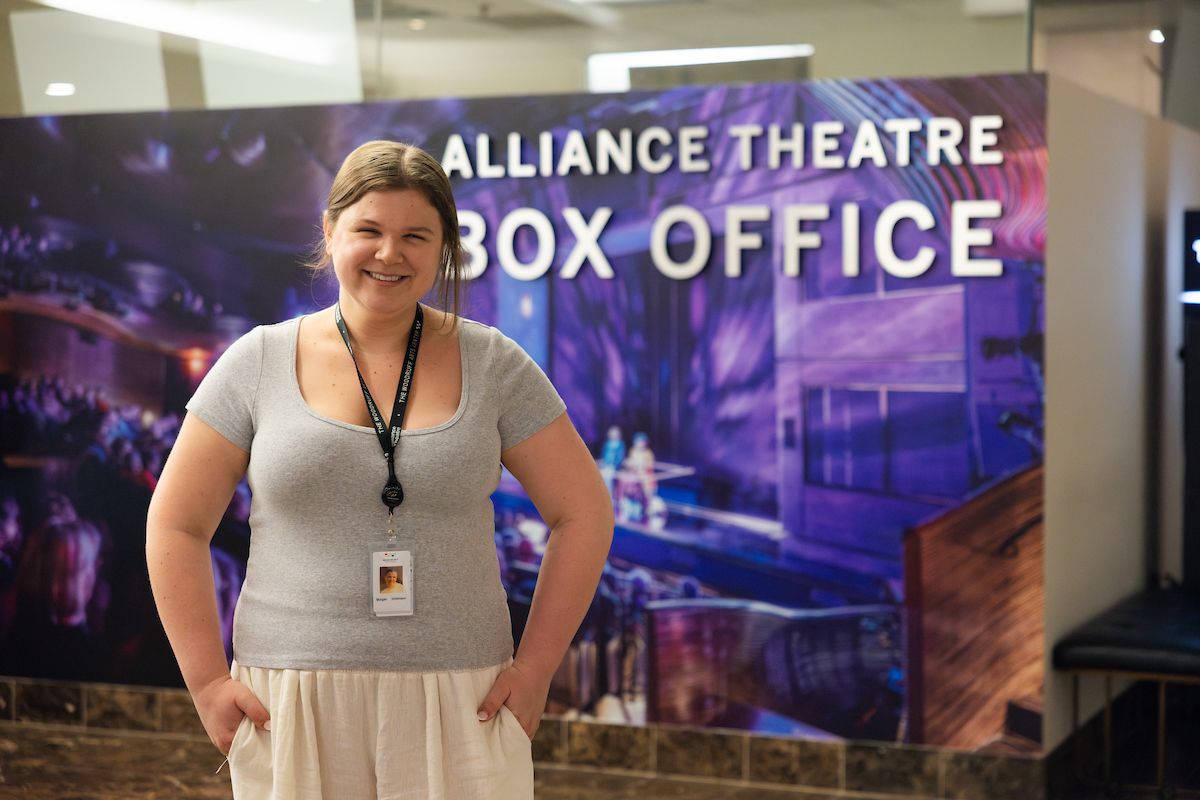 A young woman stands smiling in front of a sign that reads "Alliance Theatre Box Office."