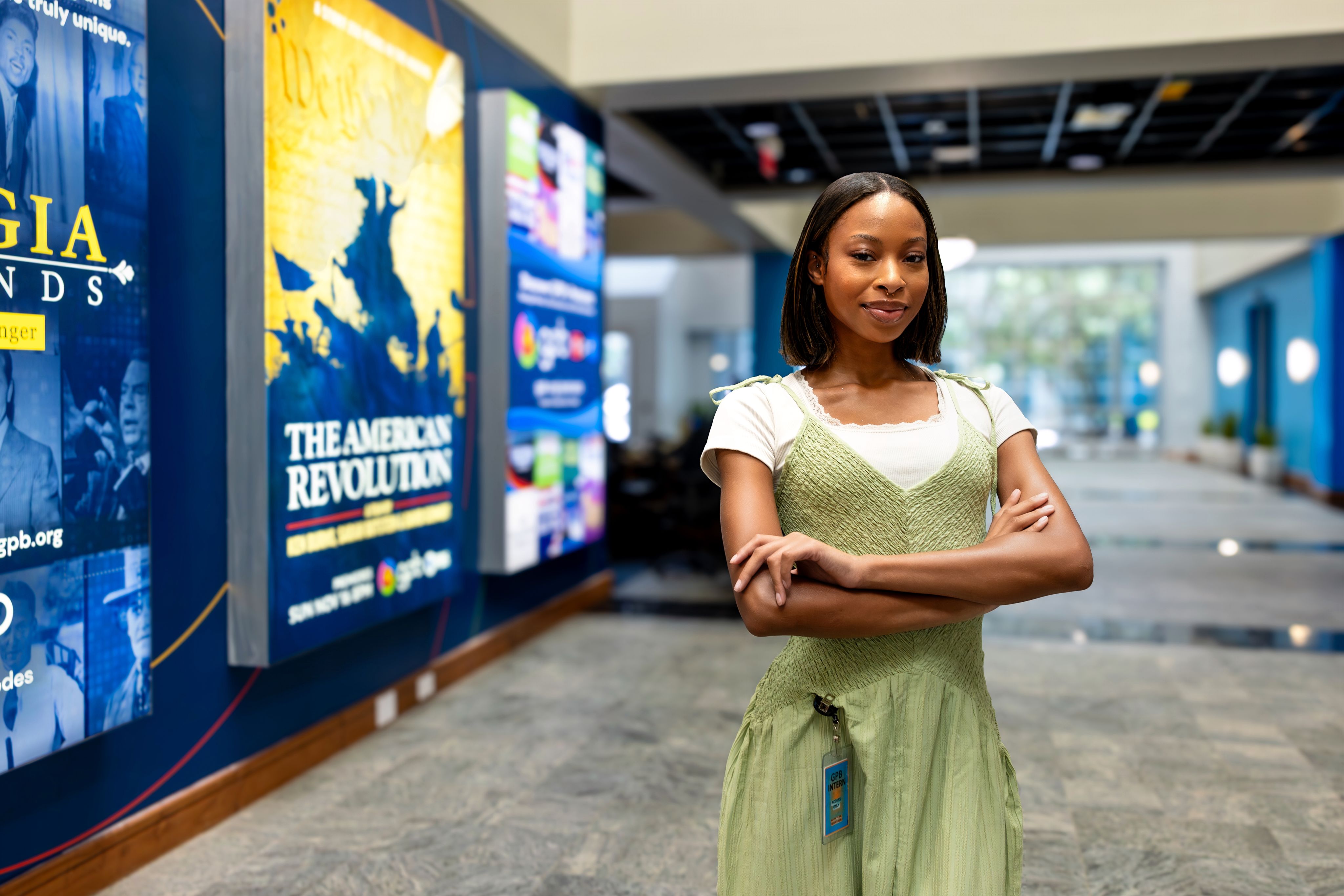 A young woman stands with her arms crossed in the hallway of a public media station. Behind her are posters advertising a variety of public broadcasting television programs.