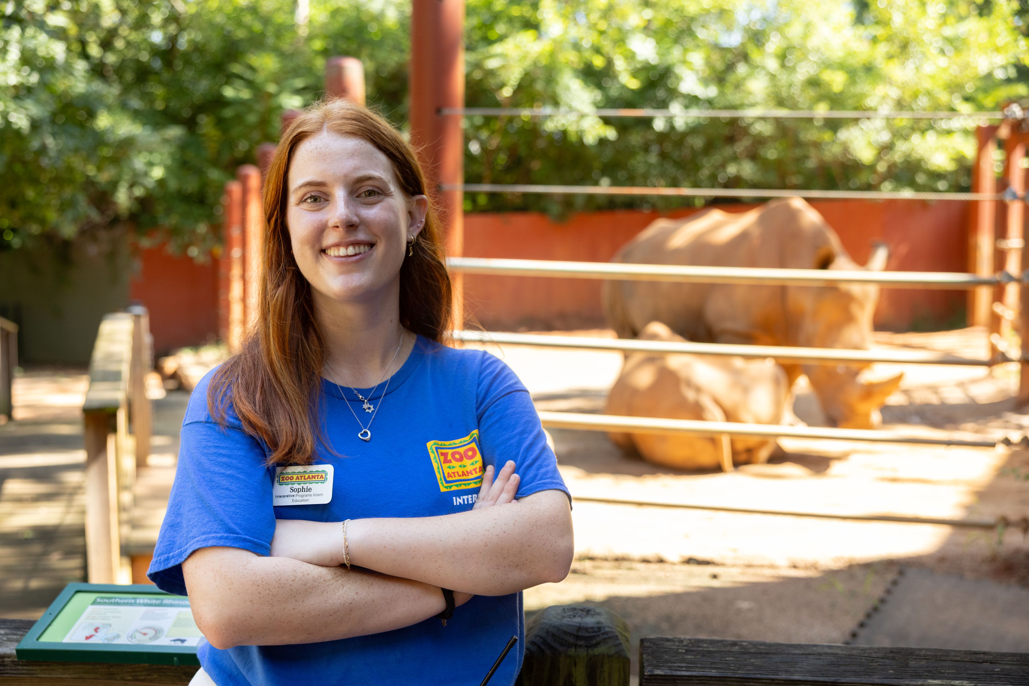 Above, a child with long, red hair, wearing a plaid skirt, poses, smiling, with her hand on the head of a white rhinoceros in an indoor zoo environment. Here, the same woman, now a college student, stands, her arms confidently crossed, in front of a rhino enclosure with two rhinos in it.