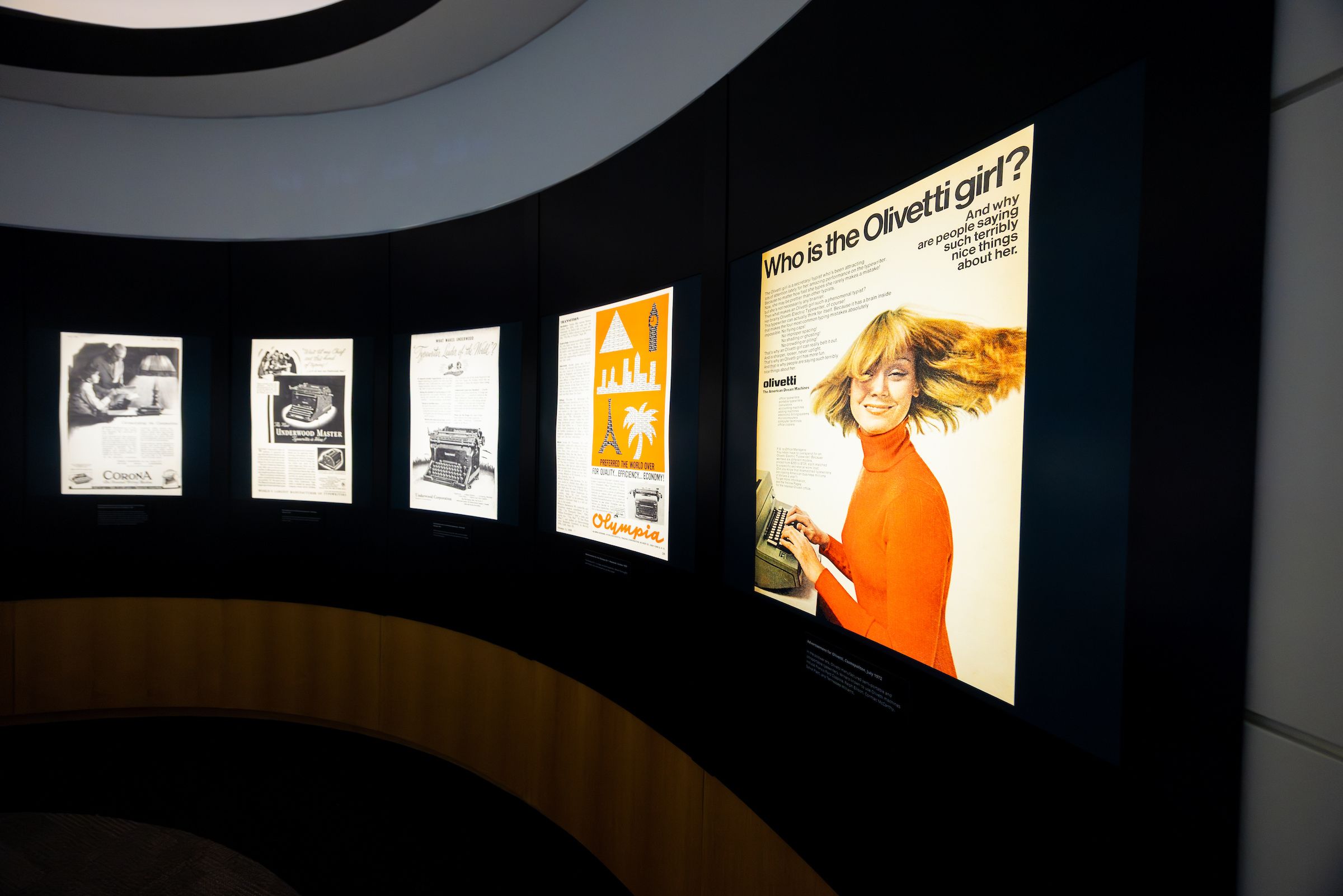 A series of five backlit vintage advertisements for typewriters, including one with a young blond white woman turning her hair so it fans out around her. Above her, the words: "Who is the Olivetti girl?"