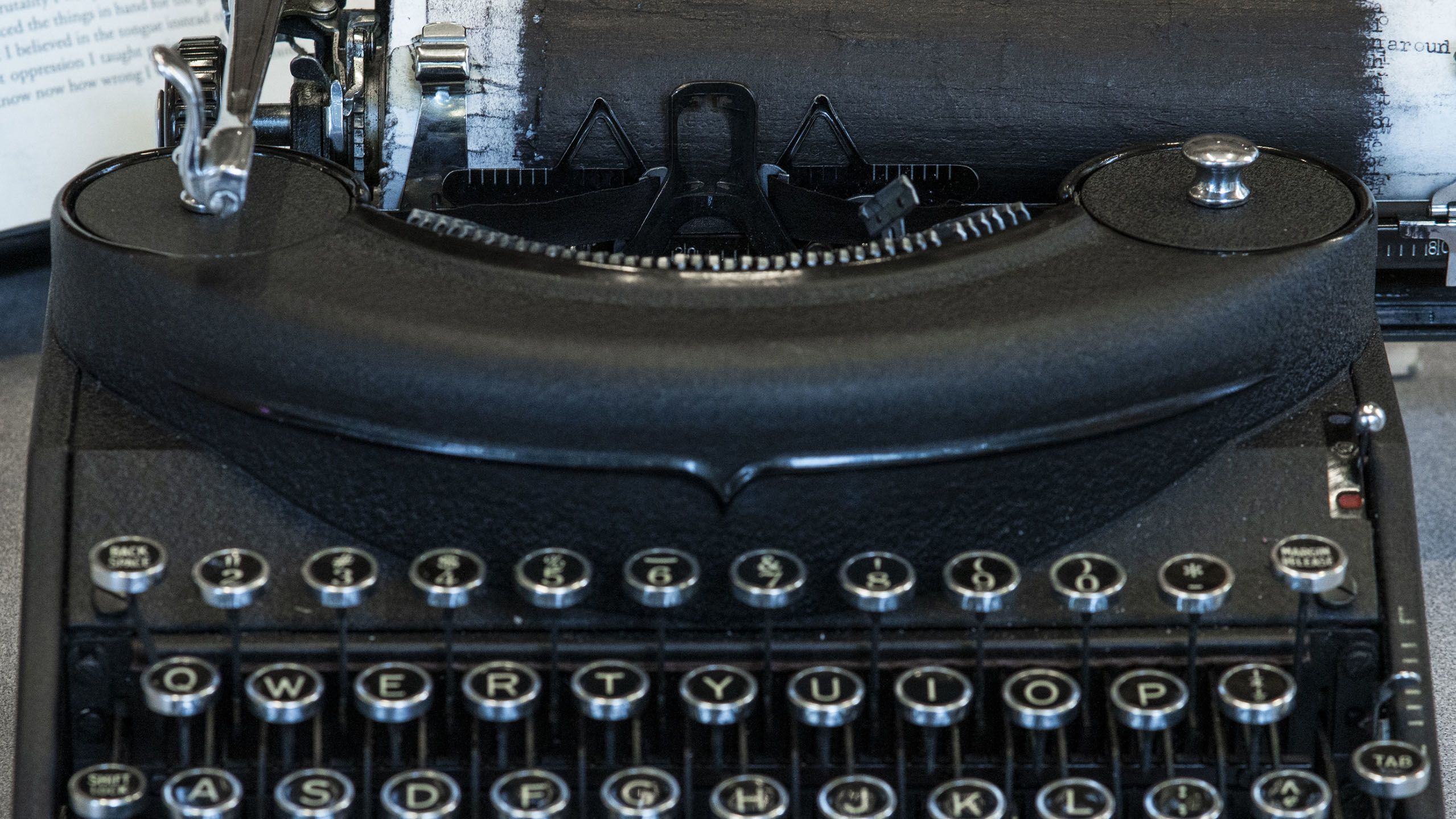 An antique typewriter sits on a table. The piece of paper it holds is covered with ink and an open book sits behind it.