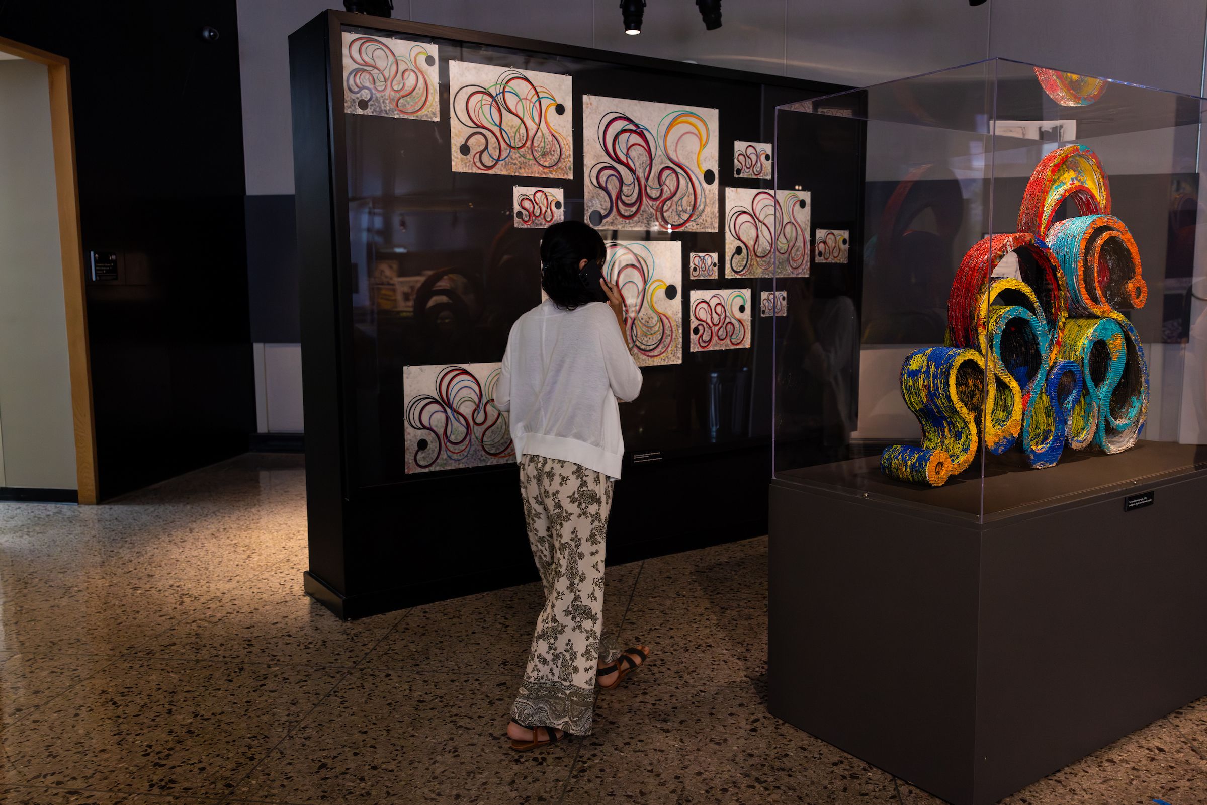 A woman walks by a series of paintings featuring colorful, looping lines. Her back it to us. Beside her, a sculpture echoing those same colorful looping lines, sits in a glass case.