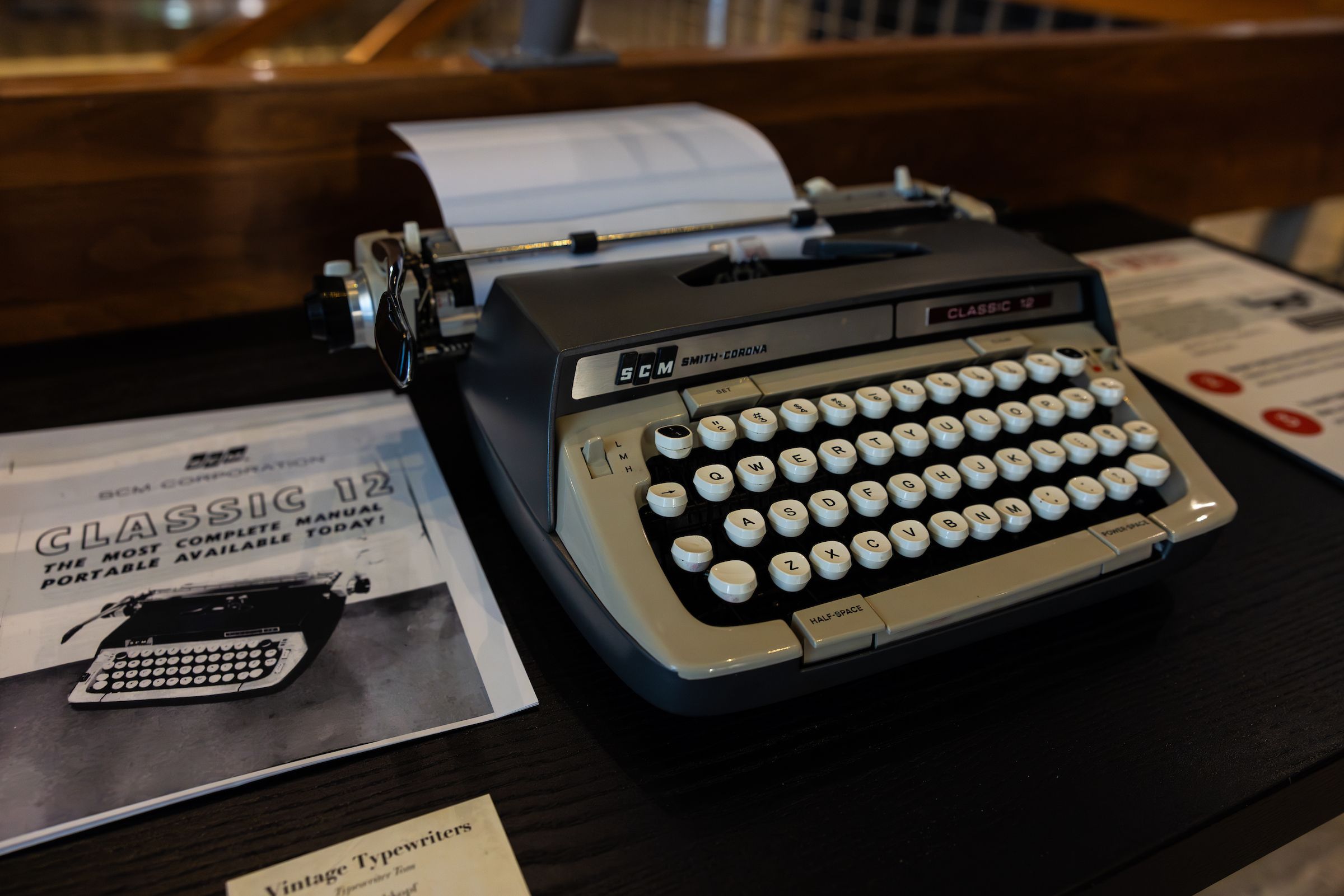 A drab green manual typewriter sits, loaded with paper, on a desk, next to its user manual.