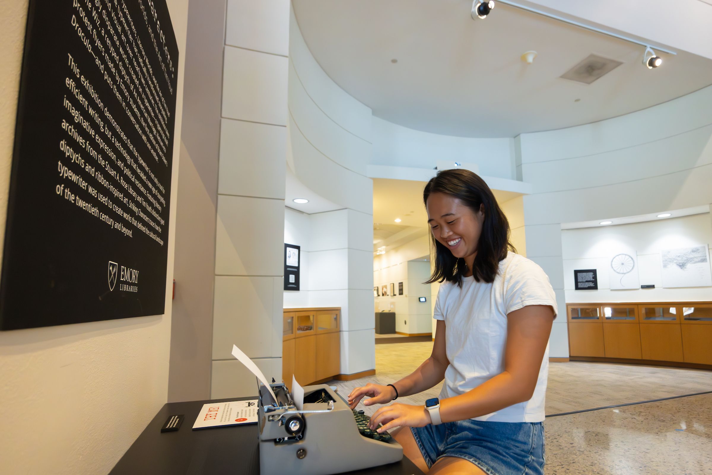 A smiling college student wearing a white top and jean shorts sits at a desk typing on a typewriter.