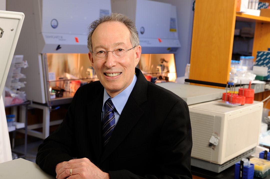 A portrait of Walter Orenstein, a white man with glasses and gray hair in a dark jacket and tie, sitting in his lab with equipment behind him.